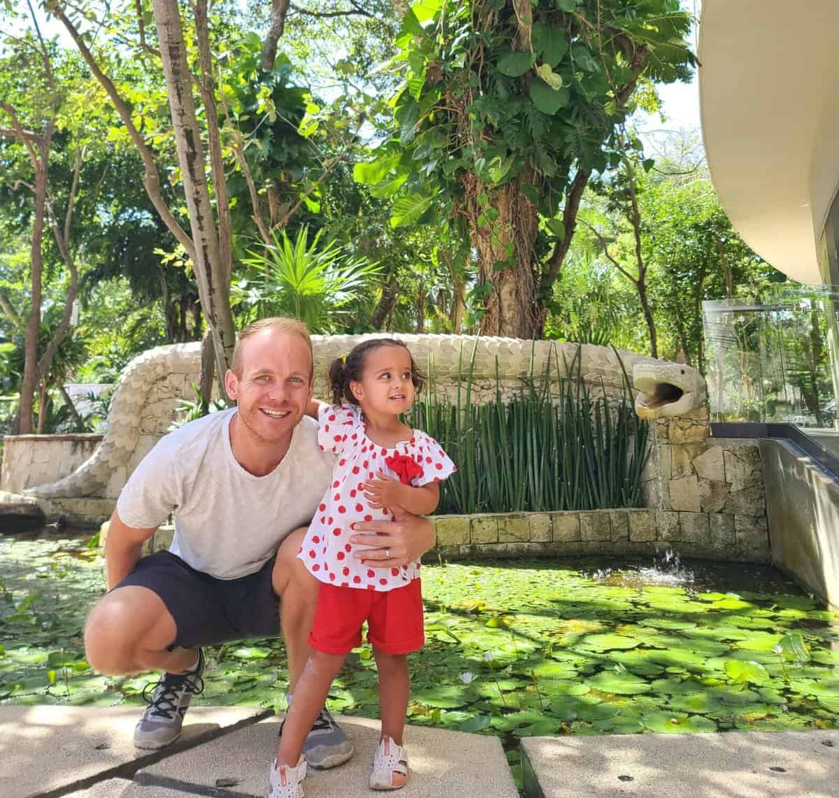 A smiling man crouching next to a young child in a white and red dress, standing by a water lily pond with a small fountain in Playa del Carmen, Riviera Maya, Quintana Roo, Mexico.