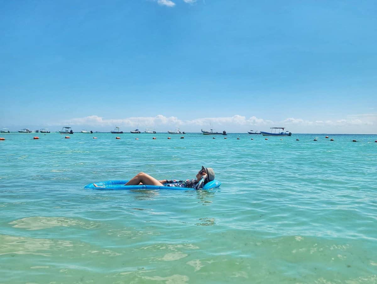 Person relaxing on a blue float in the clear, turquoise waters of Playa del Carmen, Quintana Roo, Mexico, with boats and a blue horizon in the background.