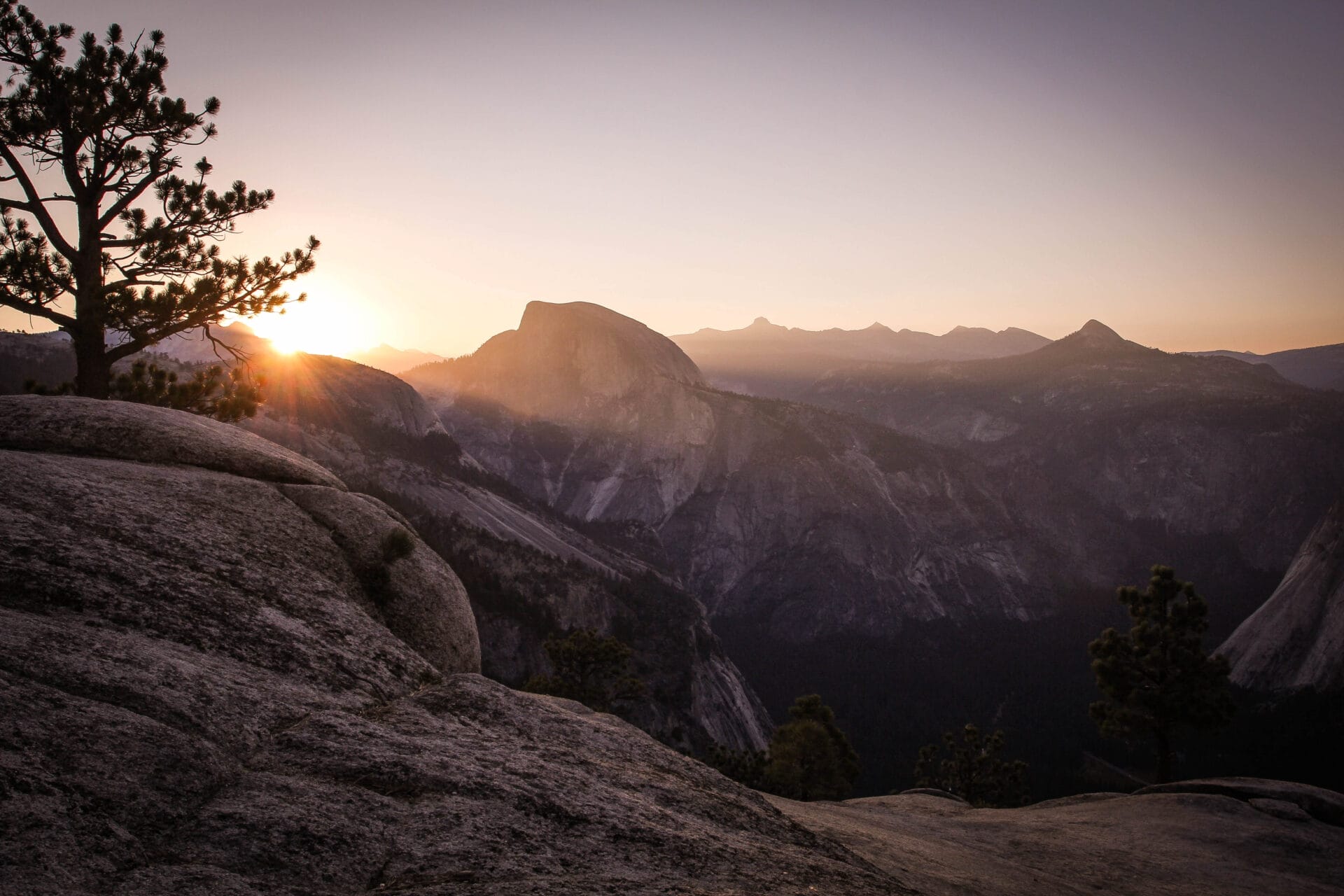 Sunset over Half Dome in Yosemite National Park with a tree silhouetted against the sun and rocky terrain in the foreground.
