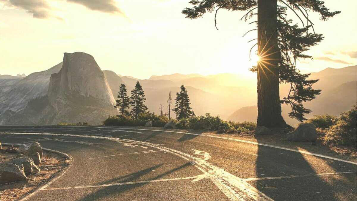 A scenic view of Glacier Point Road at sunrise in Yosemite National Park, capturing the sun peeking through a tall tree with the iconic Half Dome in the background.