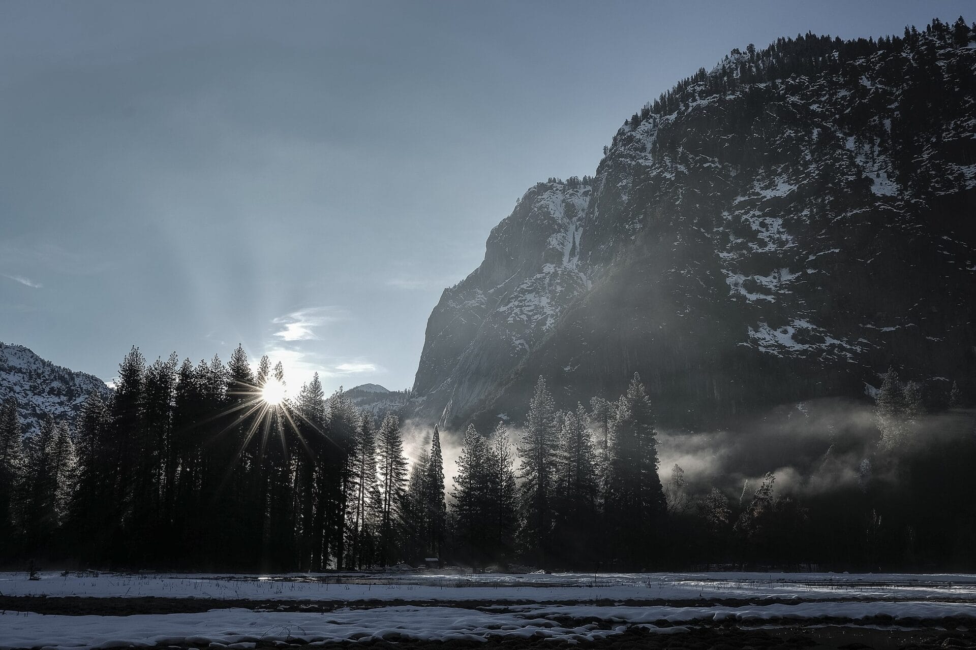 Sunrise in Yosemite National Park with sun rays shining through trees and mountains in the background.