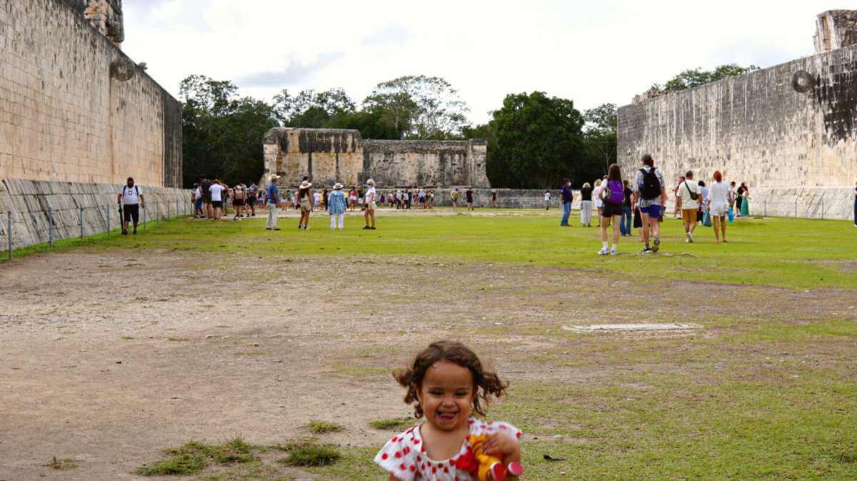 A young girl standing in the middle of a large, ancient ball court in Chichen Itza, smiling and holding a toy. The ball court is surrounded by tall stone walls and there are people walking around in the background.