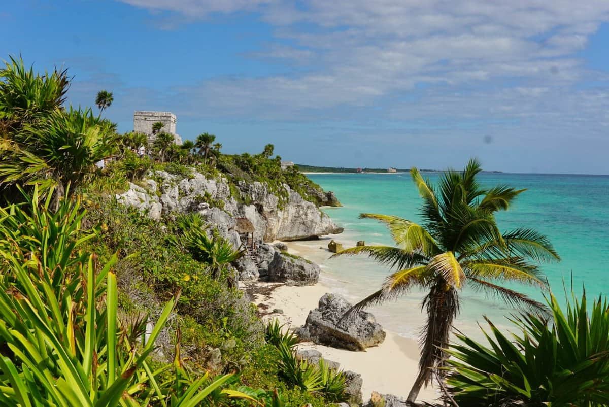 El Castillo at Tulum Ruins overlooking a sandy beach and turquoise ocean, with lush green vegetation and palm trees in Mexico.