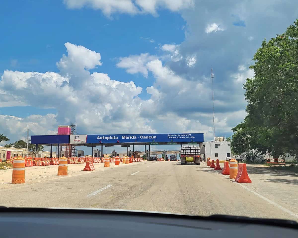 Highway toll booth on the Mérida-Cancún route in Playa del Carmen, Mexico, with traffic cones directing vehicles and a blue sky with scattered clouds overhead.
