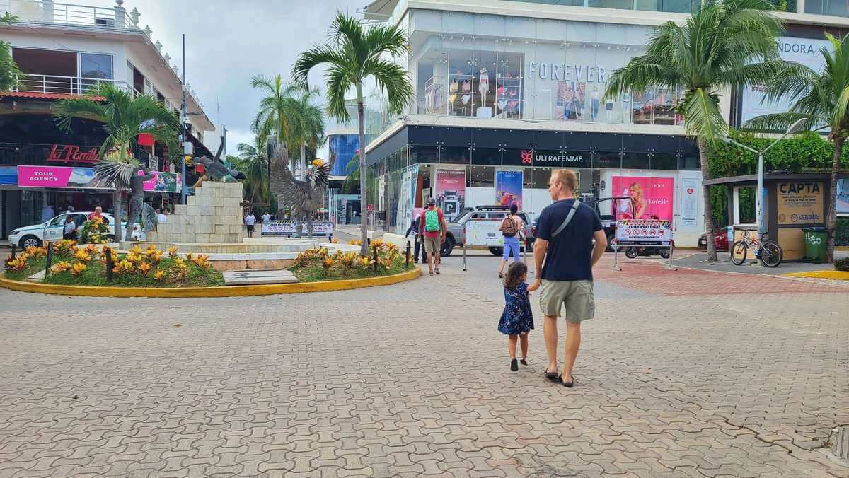 A busy street in Playa del Carmen, Riviera Maya, with people walking, shops, and a palm tree-lined avenue with a sculptured eagle centerpiece.