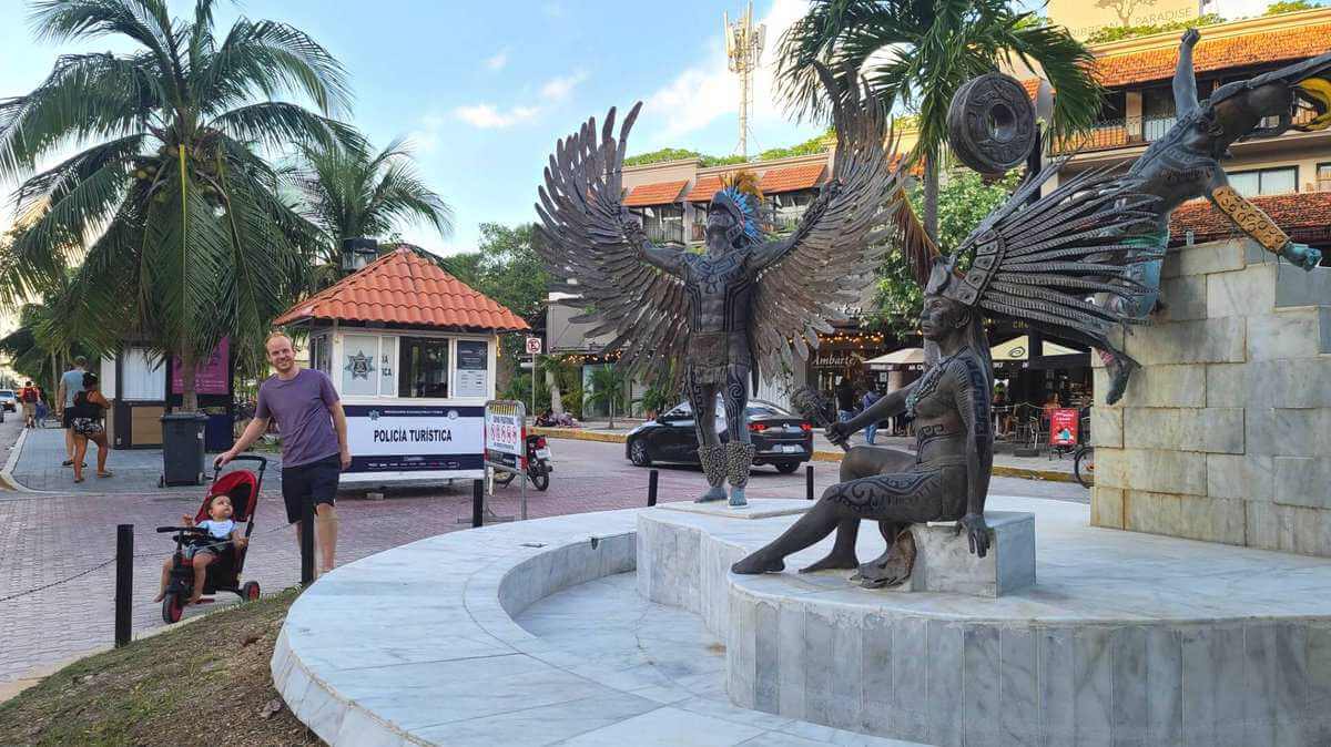 A man pushing a baby in a stroller on 5th Avenue in Playa del Carmen, Quintana Roo, Mexico. In the background, there are large bronze statues depicting indigenous dancers with feathered headdresses, and a "Policía Turística" booth is visible.