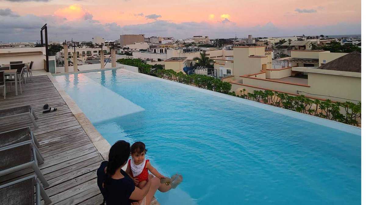 A woman and a child sit by the edge of a rooftop infinity pool with views of Playa del Carmen, Quintana Roo, Mexico in the background during sunset.