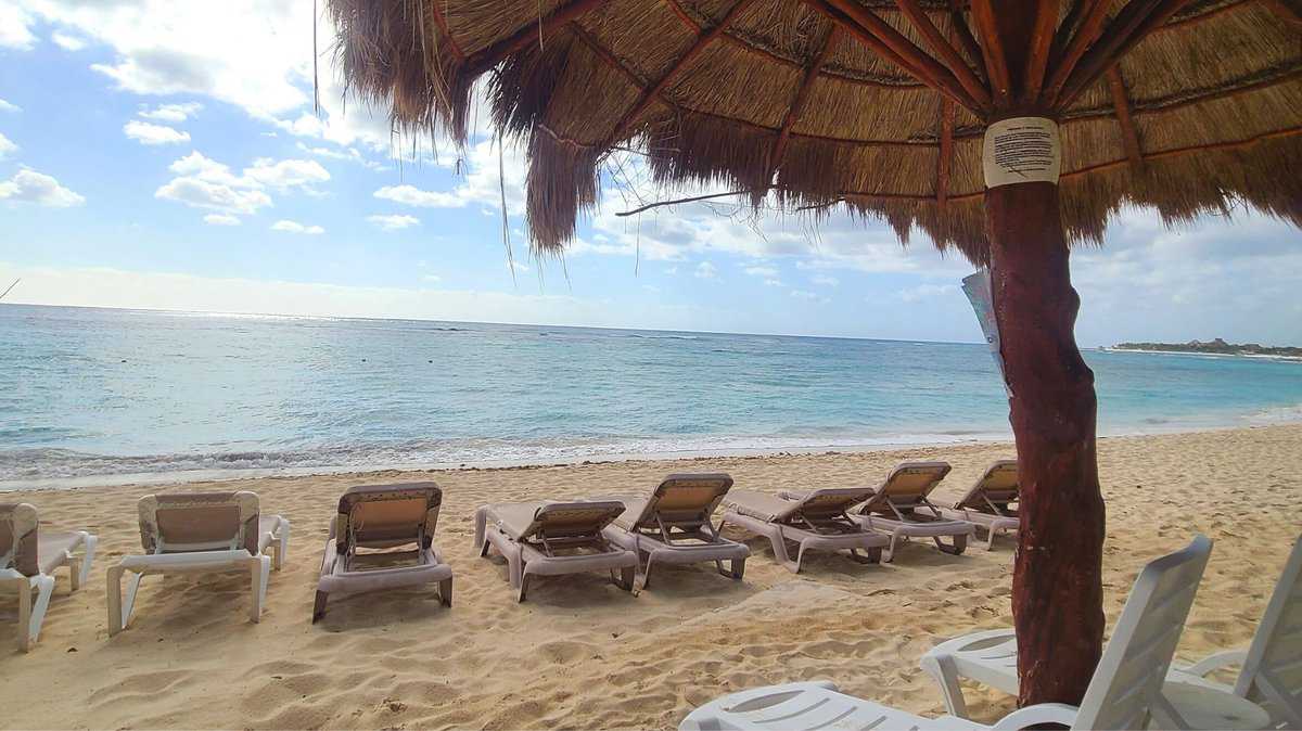 Beach chairs under a thatched-roof umbrella on a sandy beach with clear blue water in Akumal, Mexico.