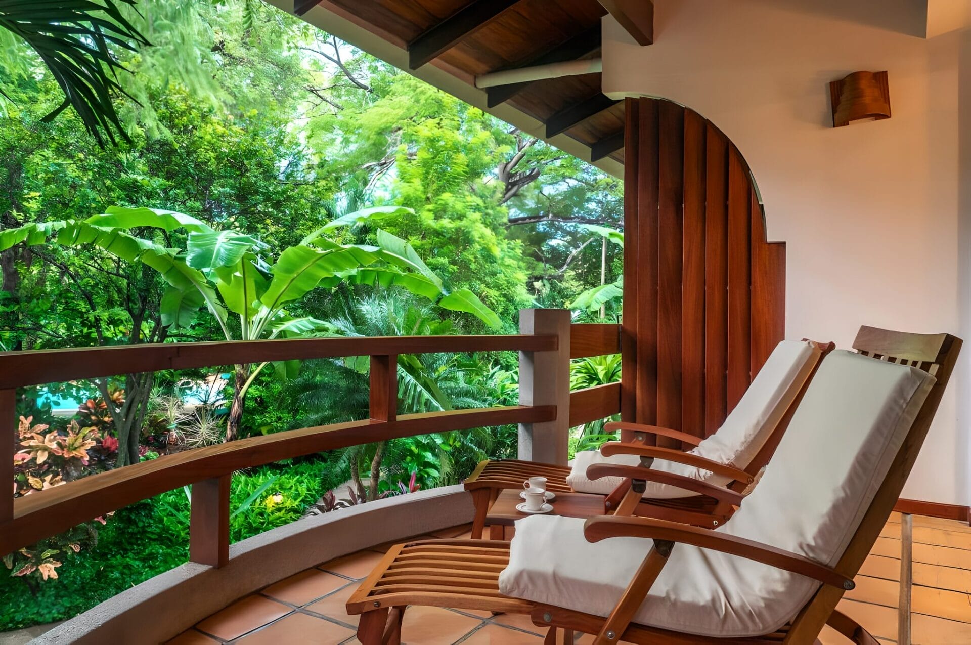 Two wooden lounge chairs with white cushions on a balcony surrounded by lush greenery at Capitán Suizo Beachfront Boutique Hotel in Tamarindo, Costa Rica.