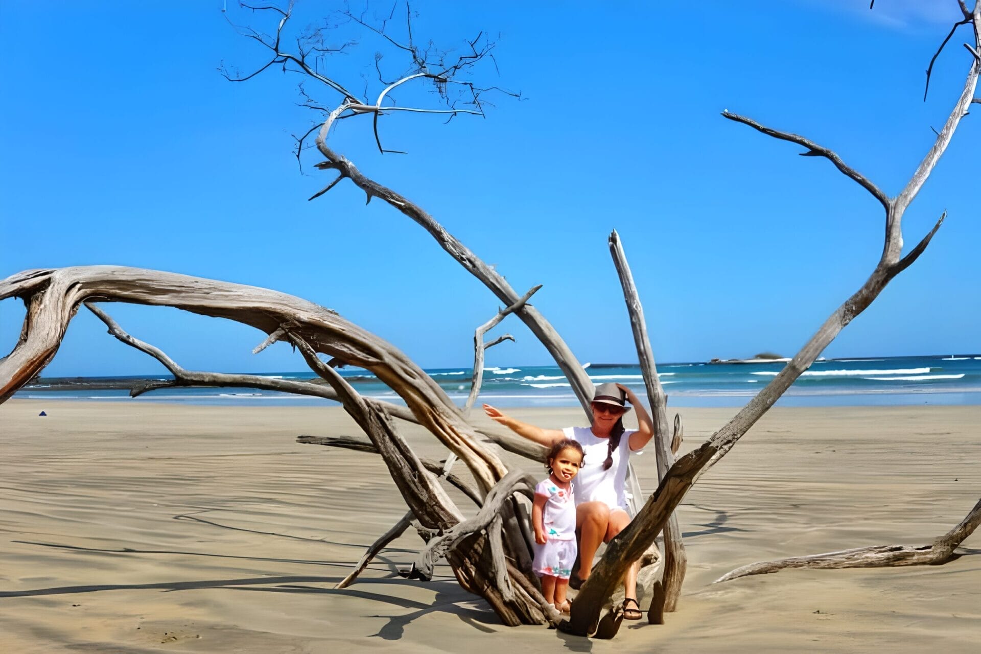 A person and a child sitting on a piece of driftwood on the beach in Tamarindo, Costa Rica. The beach has golden sand and clear blue skies, with ocean waves gently rolling in the background.