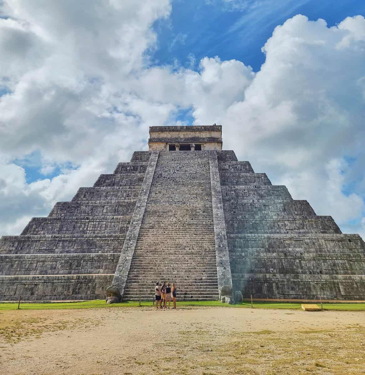 Visitors standing in front of the iconic El Castillo pyramid at Chichen Itza under a cloudy blue sky in Yucatan, Mexico.