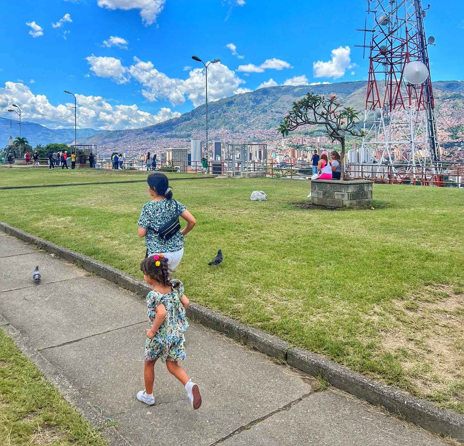 Children running on a path at Pueblito Paisa in Medellin, Colombia, with a scenic view of mountains and the cityscape in the background.