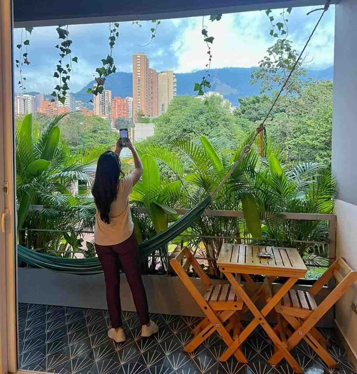 A person taking a photo from a balcony at an Airbnb in El Poblado, Medellin, Colombia. The balcony is decorated with plants, a hammock, and a wooden table with chairs. The background shows high-rise buildings and mountains.