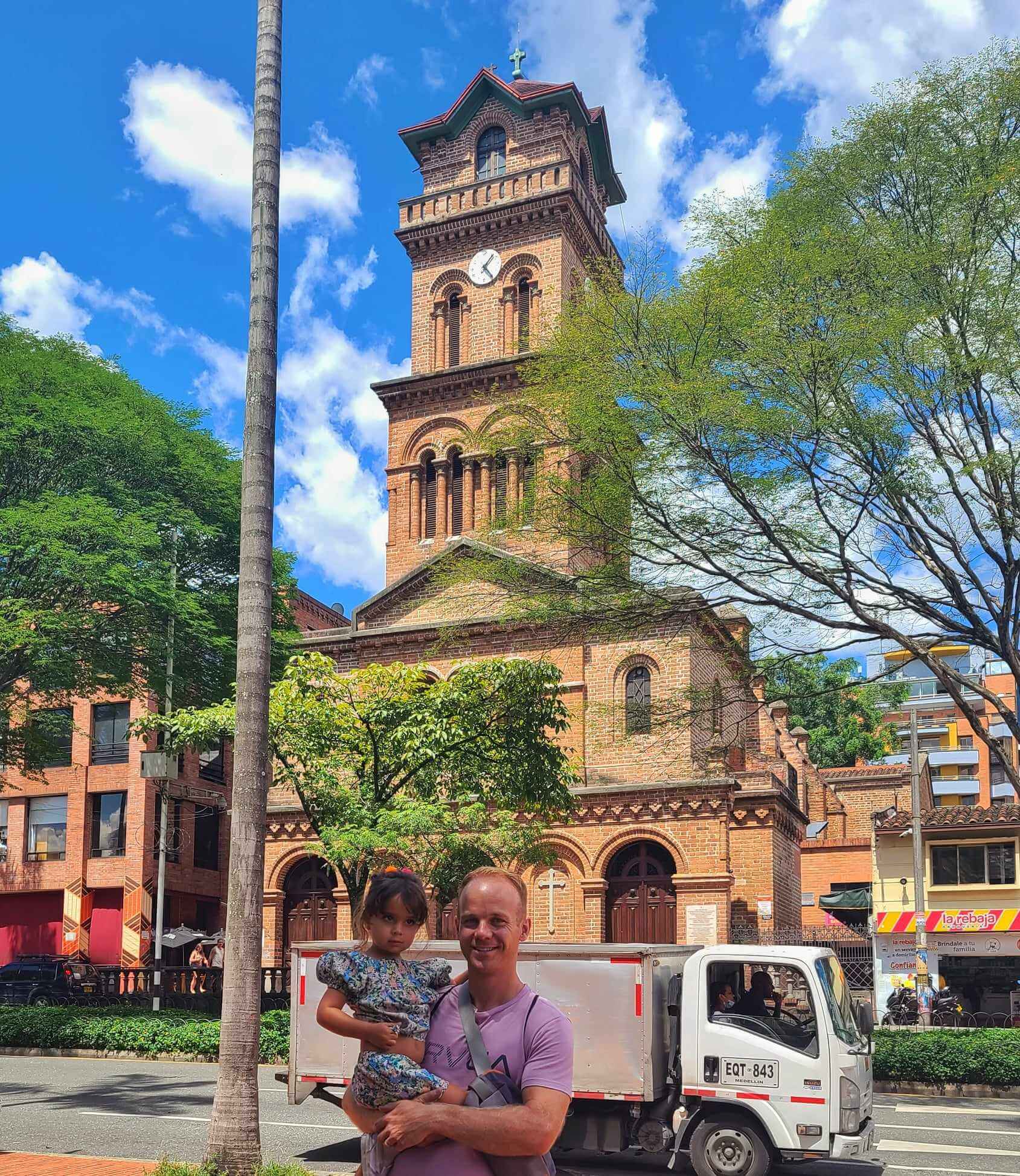 A man holding a child in front of Virgen de la Candelaria Church located in Parque De El Poblado, Medellin, Colombia, with a blue sky and clouds in the background.