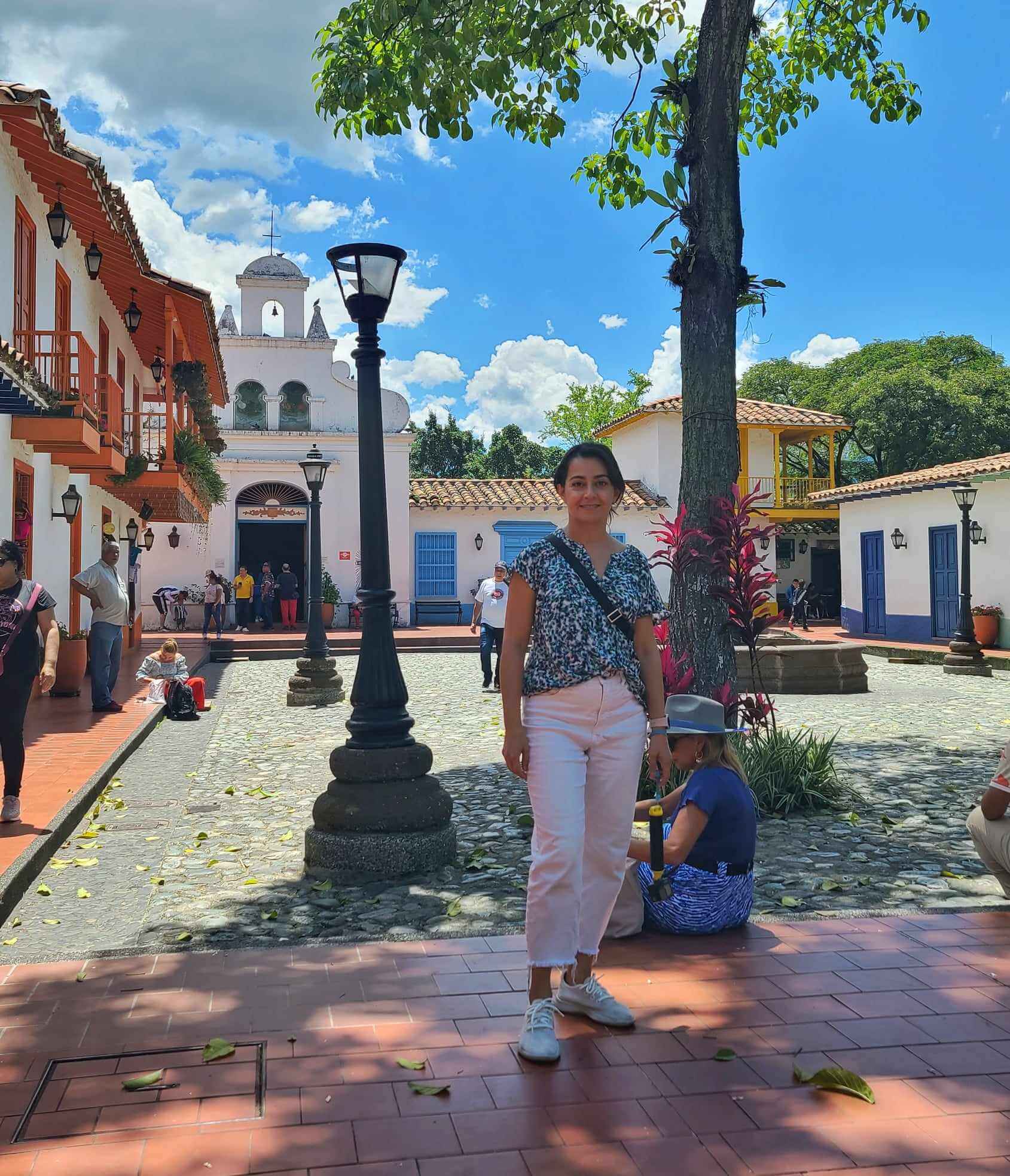A woman stands in the foreground of Pueblito Paisa in Medellin, Colombia. She is surrounded by colorful colonial-style buildings, cobblestone paths, and a few other people walking and sitting in the plaza. A white church with a bell tower is visible in the background, beneath a bright blue sky with scattered clouds.