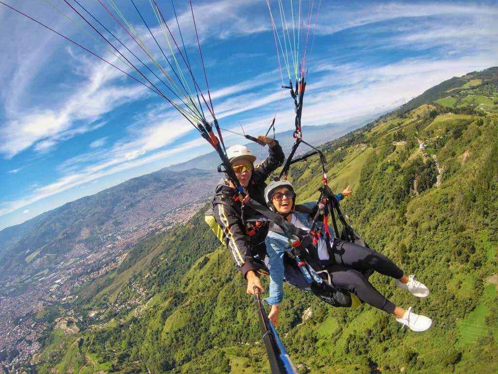 A person and an instructor paragliding over the lush green hills of Medellin, Colombia, with a panoramic view of the city and mountains in the background.