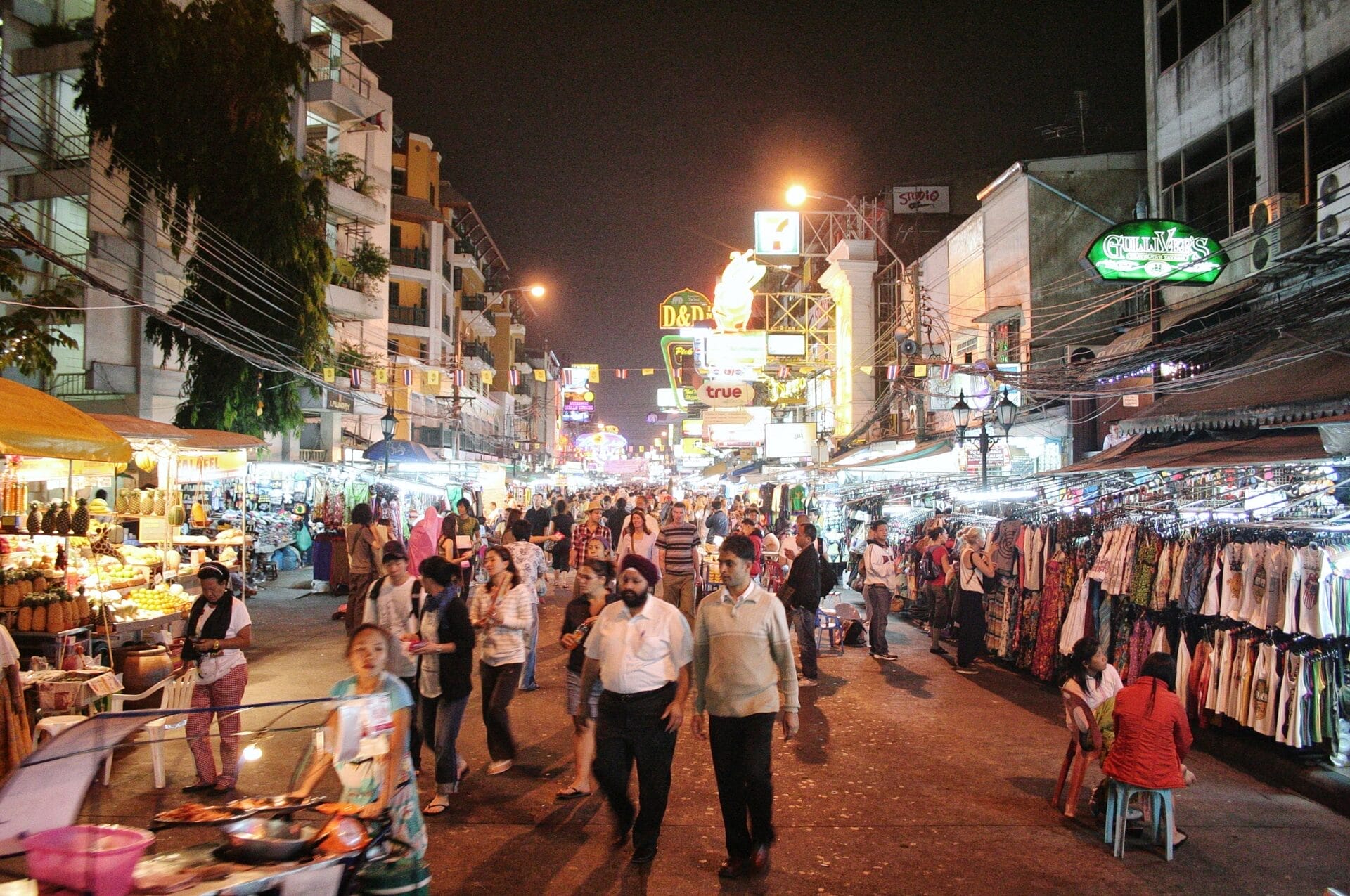 A vibrant street scene at night on Khaosan Road in Bangkok, filled with people walking past various street stalls and illuminated signs.