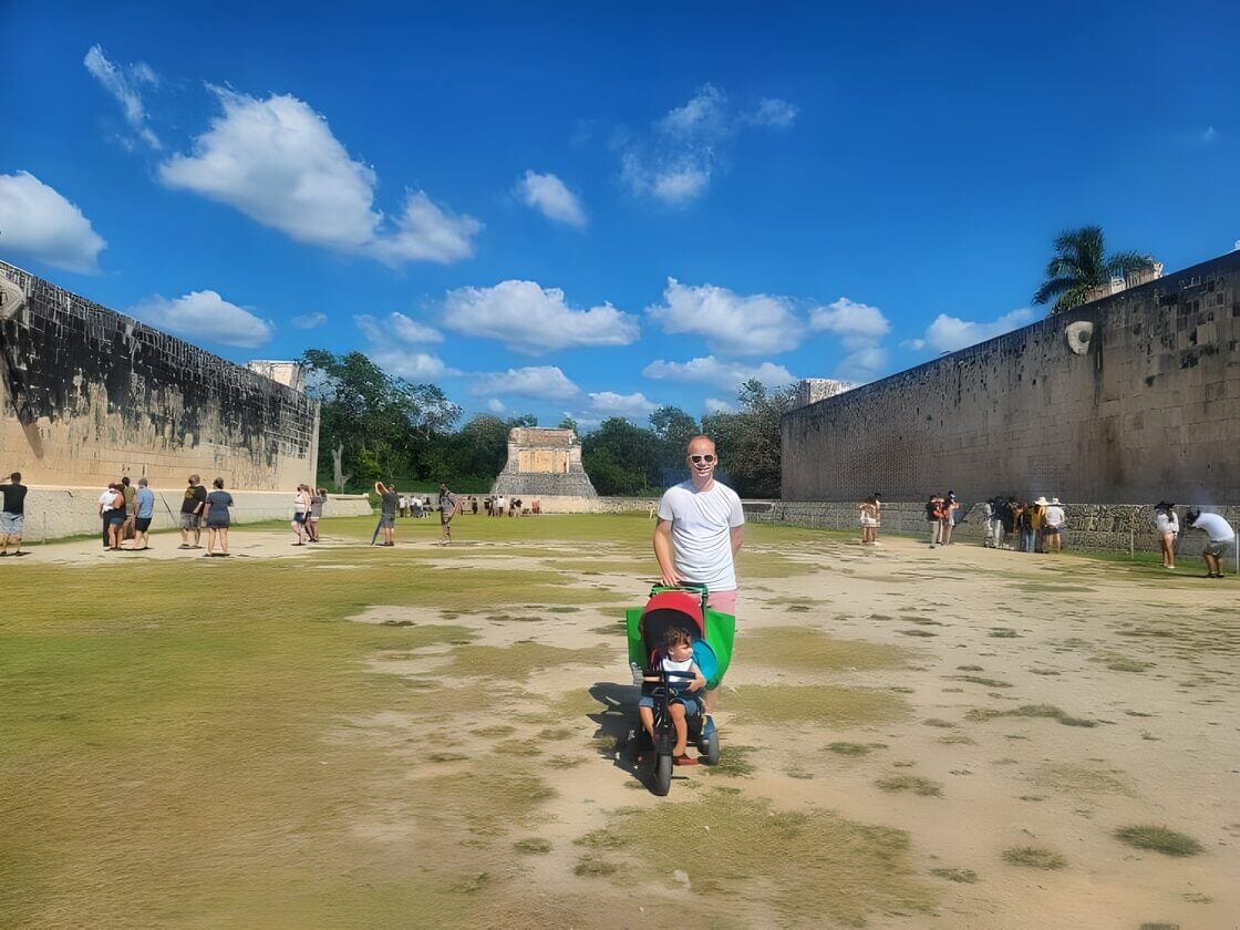 A man smiling while pushing a child in a stroller at the ancient ball court in Chichen Itza, with other tourists and clear blue sky in the background.
