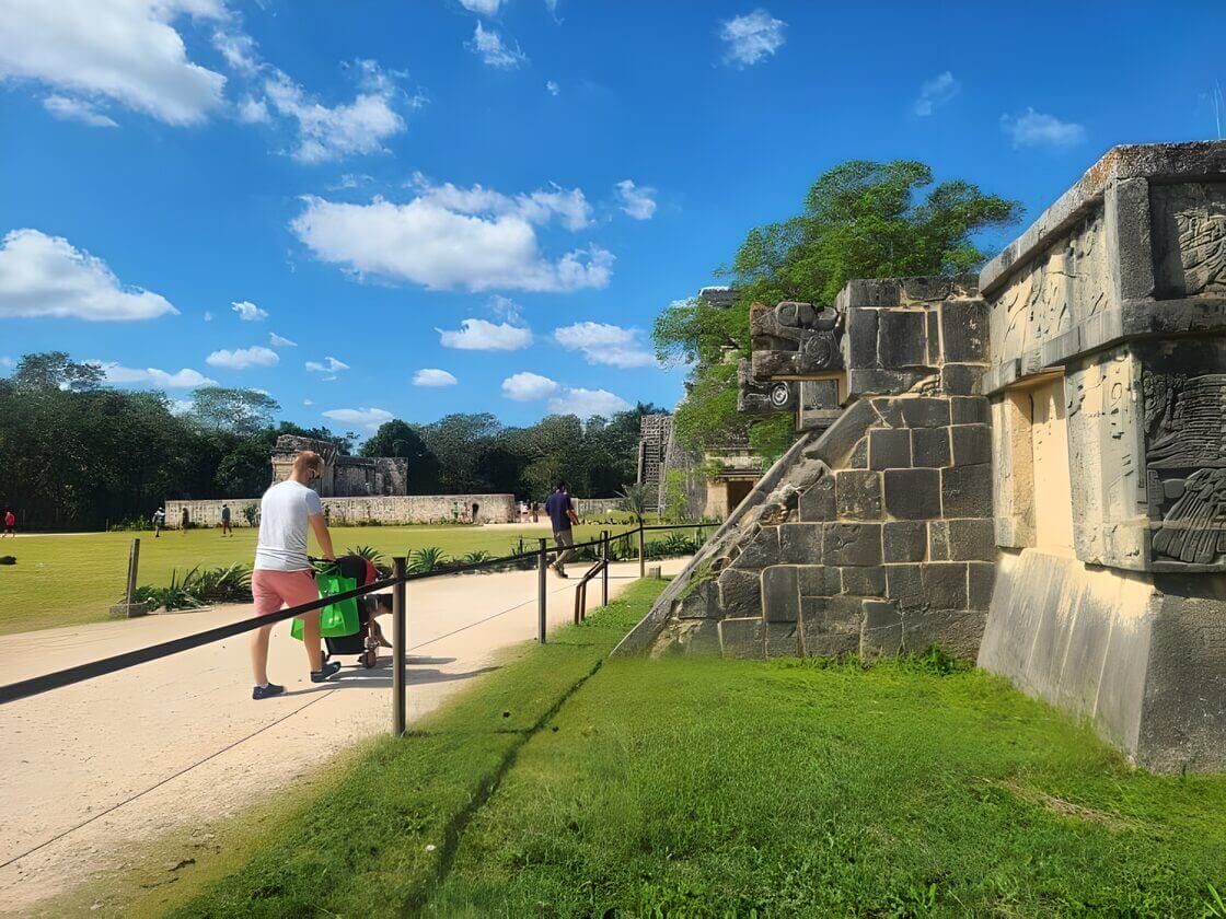A man pushing a stroller walks past a large stone structure with intricate carvings in Chichén-Itzá, Mexico.
