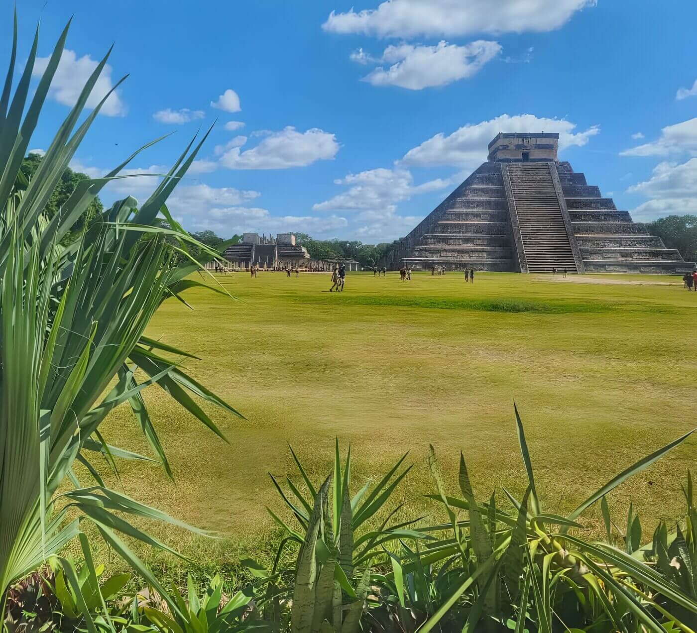 A scenic view of El Castillo, the Temple of Kukulcan, at Chichen Itza with a bright blue sky and tourists exploring the grounds, framed by lush green foliage in the foreground.