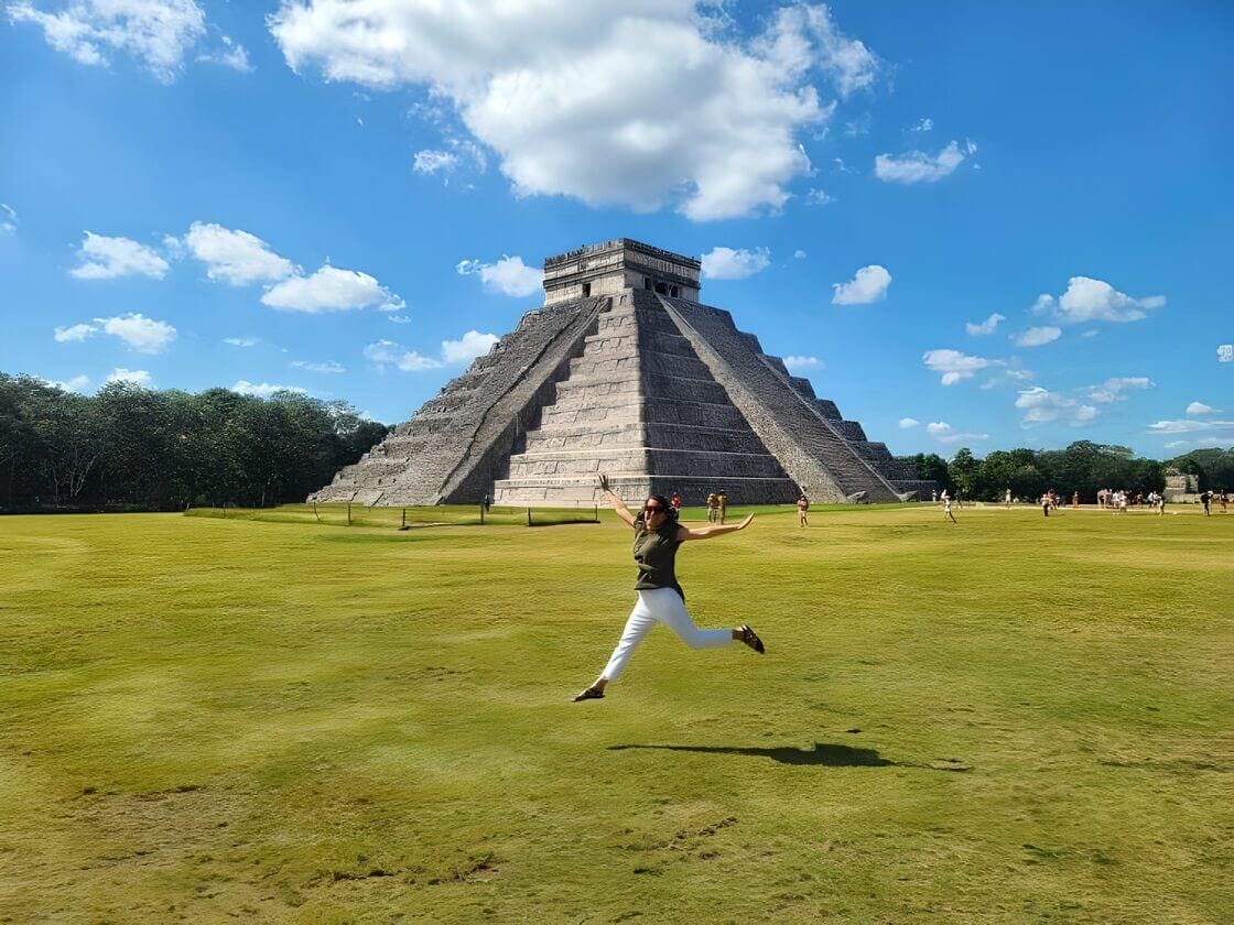 Woman jumping in front of the ancient Mayan pyramid of Chichen Itza in Yucatan, Mexico on a sunny day with blue skies and scattered clouds.