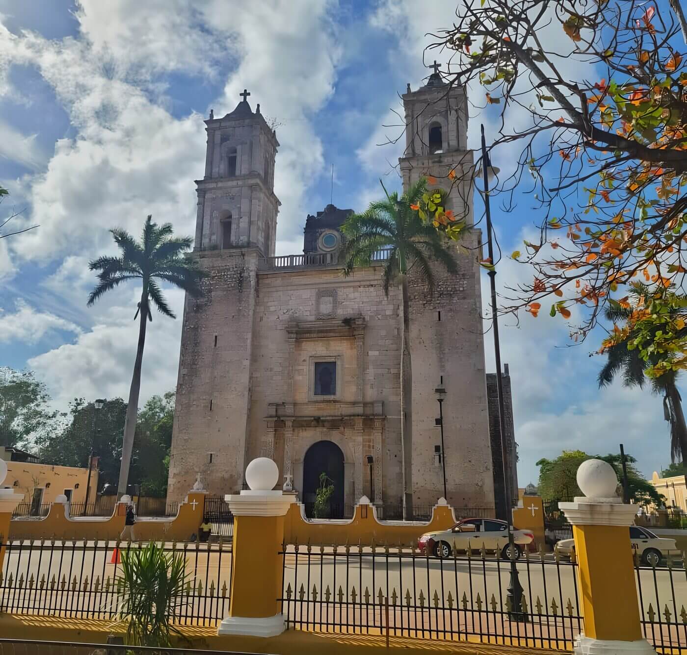 A photo of the facade of the San Gervasio Cathedral in Valladolid, Yucatan, Mexico. The cathedral is made of white stone and has two bell towers. There are palm trees in front of the cathedral and a blue sky with white clouds in the background.