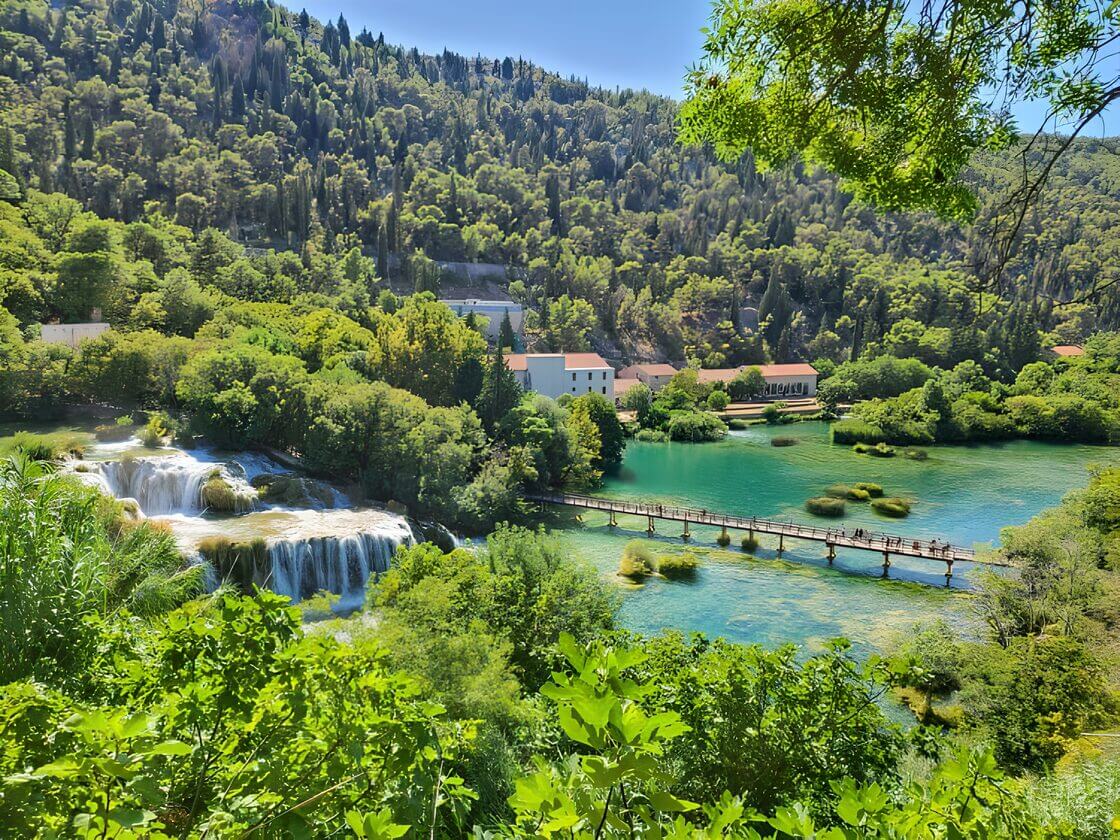 Croatia Nationa Park with kids-Krka waterfall View of Krka National Park in Croatia, featuring a stunning waterfall, a wooden bridge over clear turquoise waters, and a lush, green landscape under a bright blue sky.
