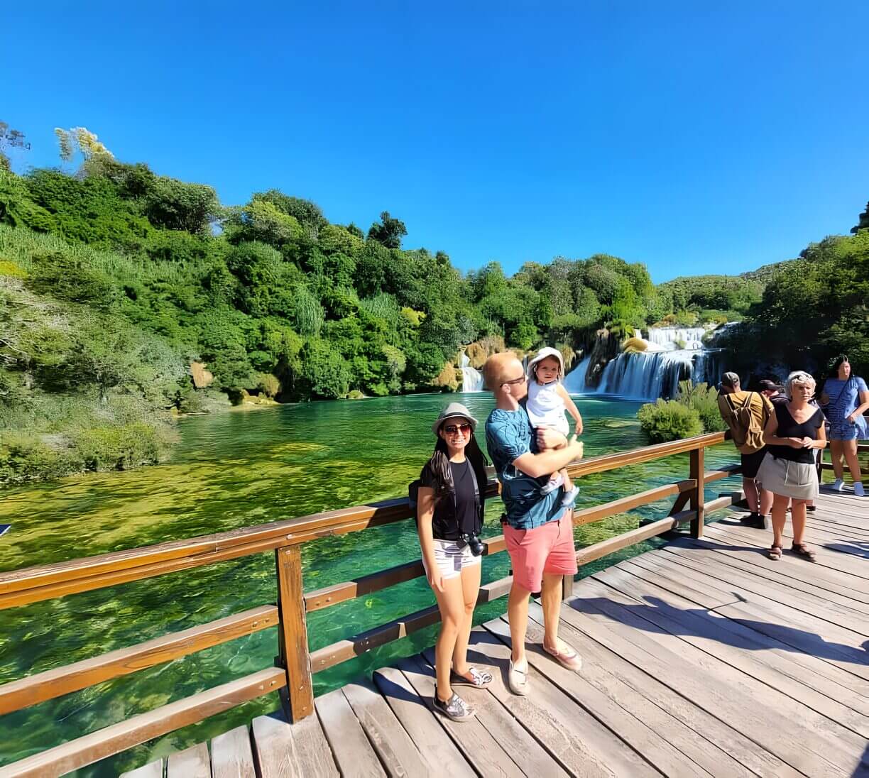 Krka main waterfall A family standing on a wooden bridge with lush greenery and a waterfall in the background at Krka National Park, Croatia.