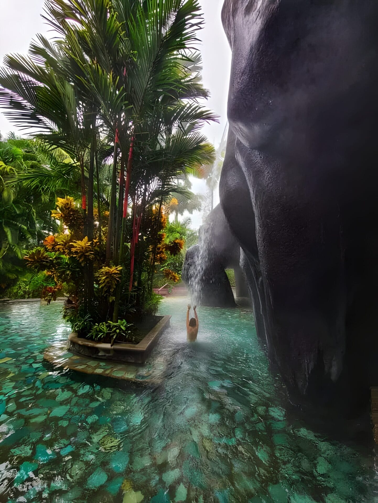 Thermal Baths in La Fortuna - Costa Rica two week itinerary Person enjoying a thermal pool with a waterfall in La Fortuna, Costa Rica surrounded by lush tropical vegetation.