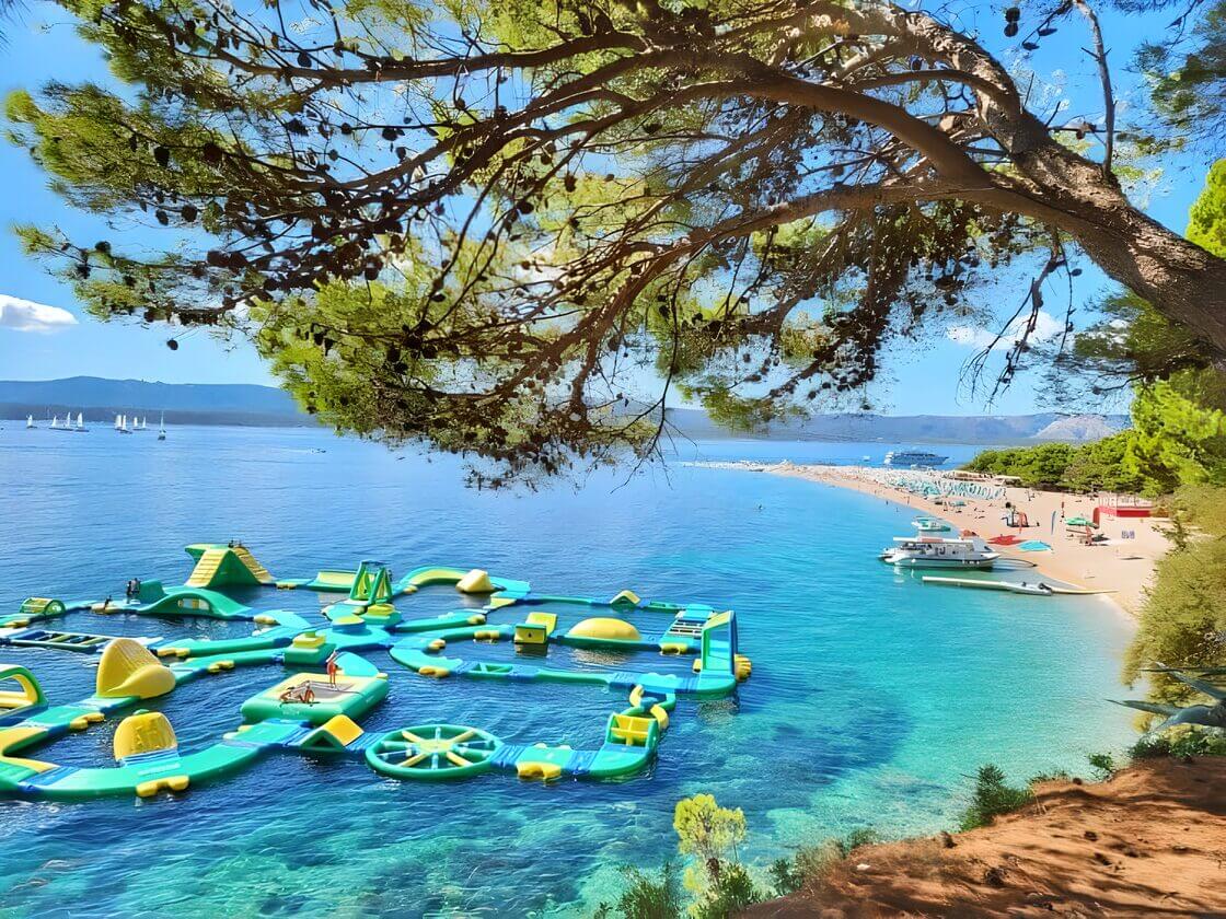 Zlatni Rat Beach - visit Croatia with kids View of Zlatni Rat beach in Bol, Brac, Croatia with clear turquoise water and an inflatable water park. The beach is lined with pine trees and boats docked along the shore.