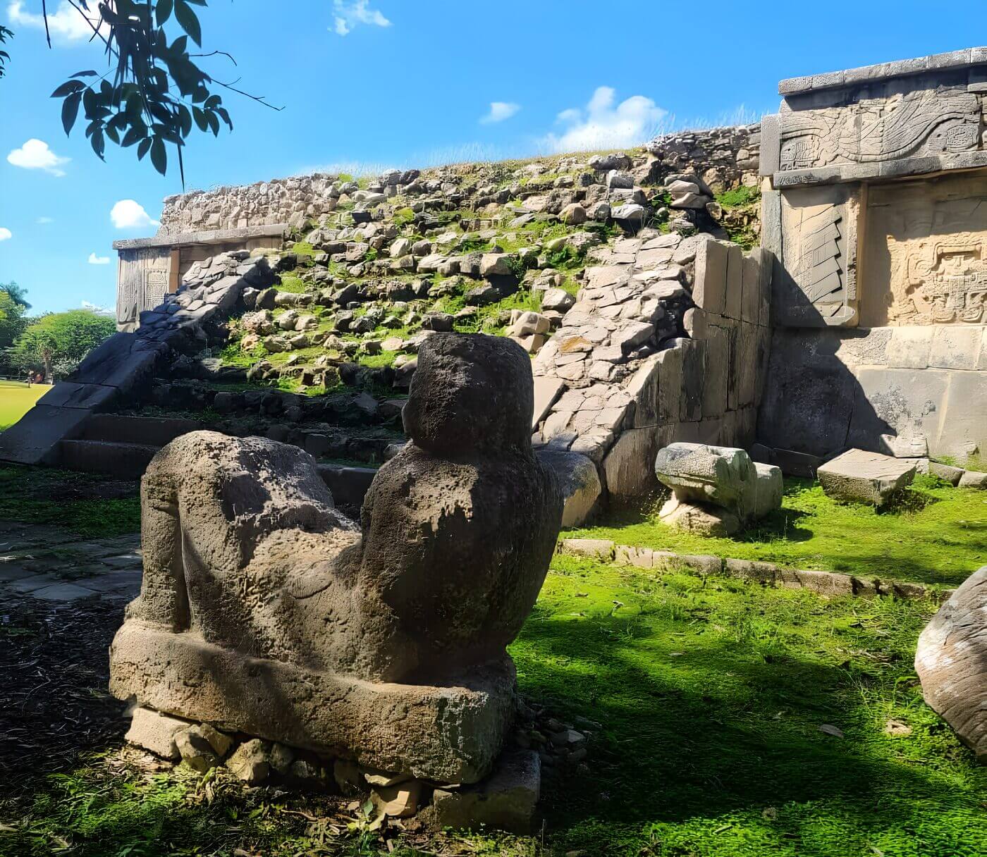 A large stone carving at the base of a partially collapsed ancient pyramid structure in Chichen Itza, Yucatan, Mexico, with green vegetation growing between the stones under a clear blue sky.
