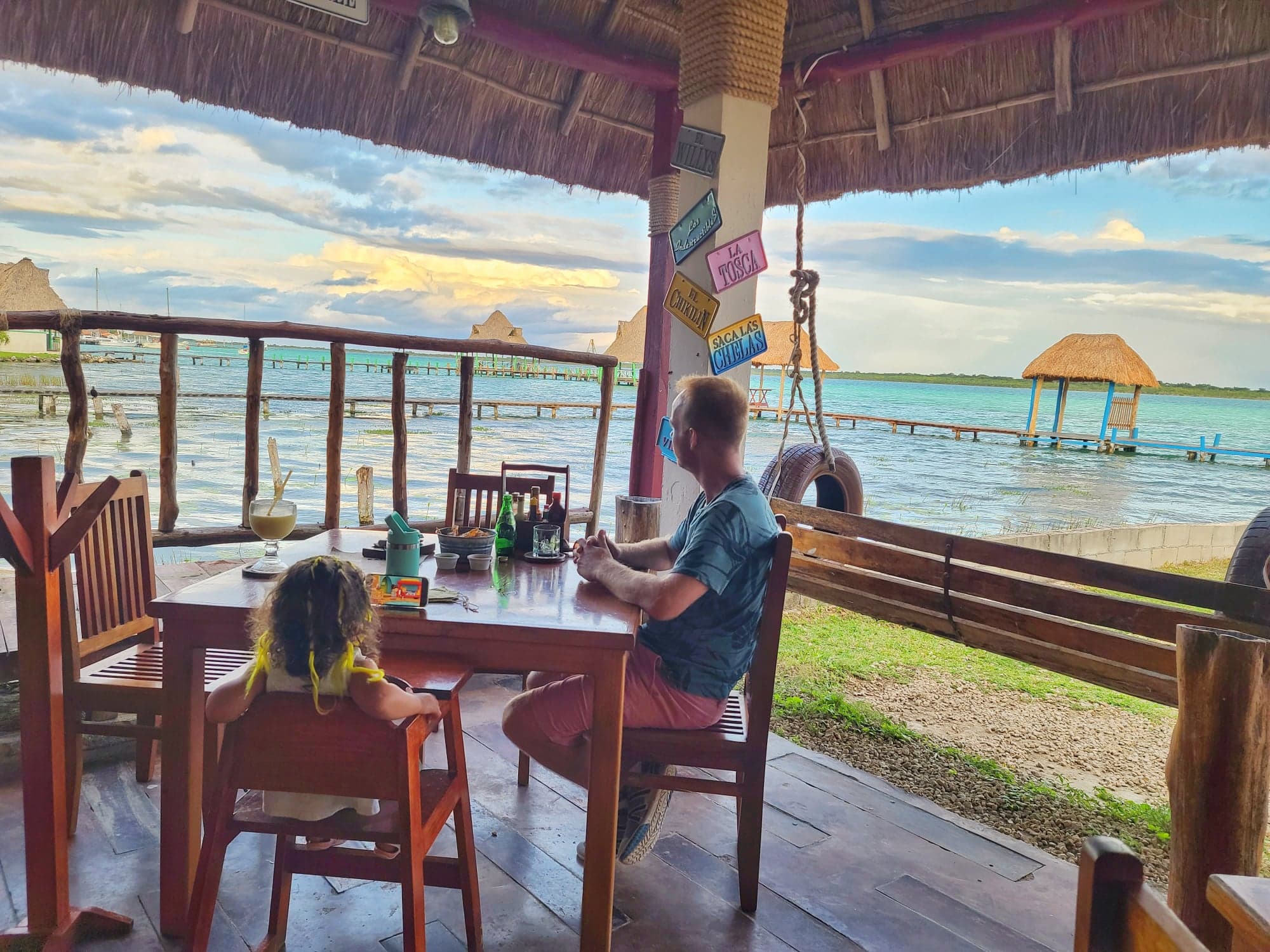Bacalar Things to do with family A picturesque view from a lakeside restaurant in Bacalar, Quintana Roo, Mexico, with a man and a child sitting at a wooden table enjoying the scenery of the tranquil blue lake and thatched roof structures.