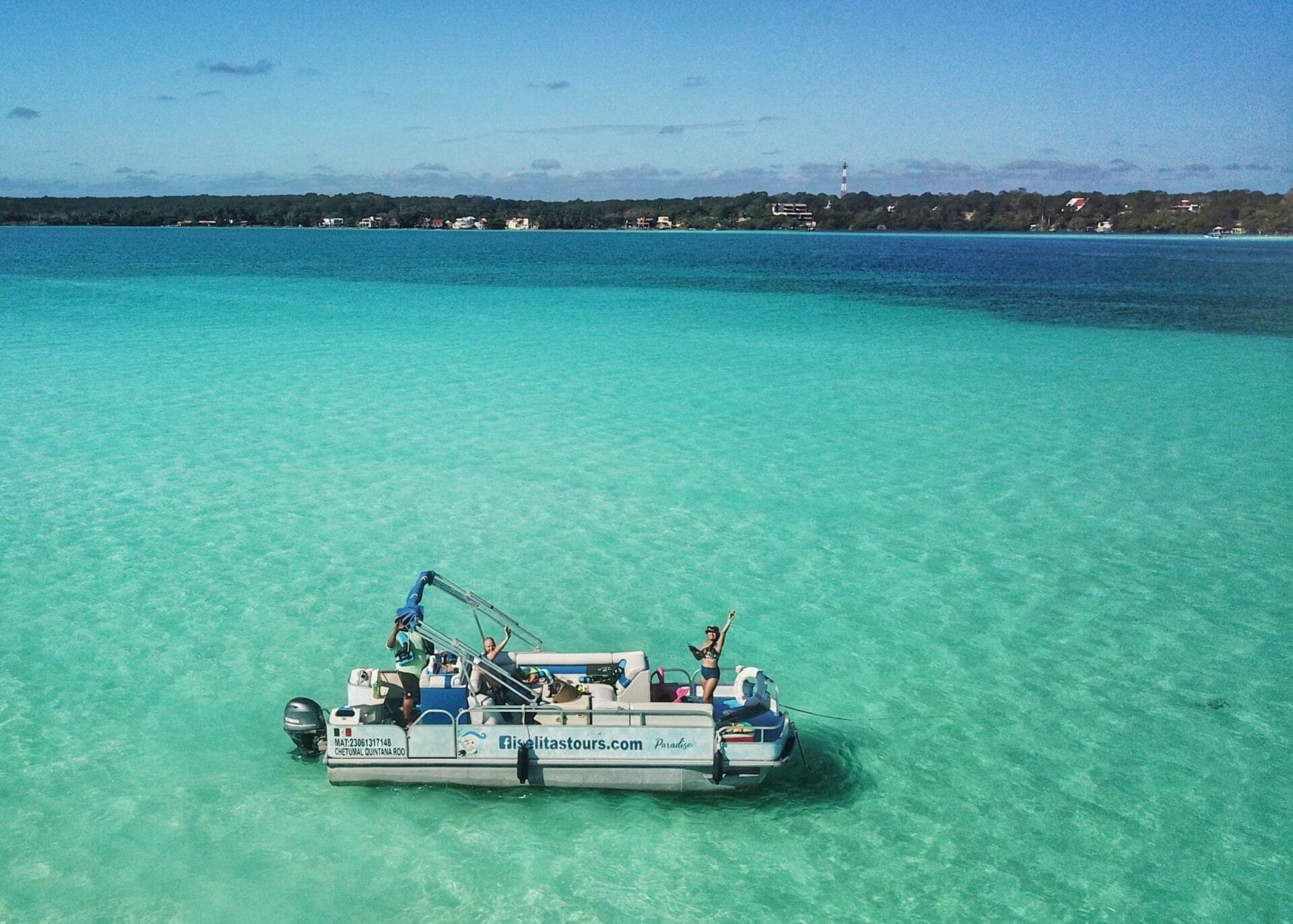 Boat tour of Bacalar Laguna A boat floating on the crystal-clear turquoise waters of Bacalar Lagoon in Quintana Roo, Mexico. The boat, labeled with "iselitastours.com," has two people on board waving and enjoying the sunny day.