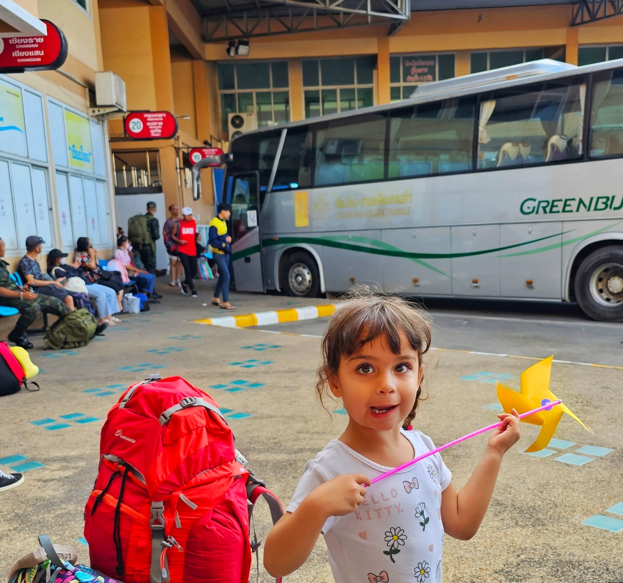 A young girl holding a toy windmill standing at a bus terminal in Chiang Mai, Thailand, with a large red backpack on the ground beside her and a group of people seated in the background near a green and white bus.
