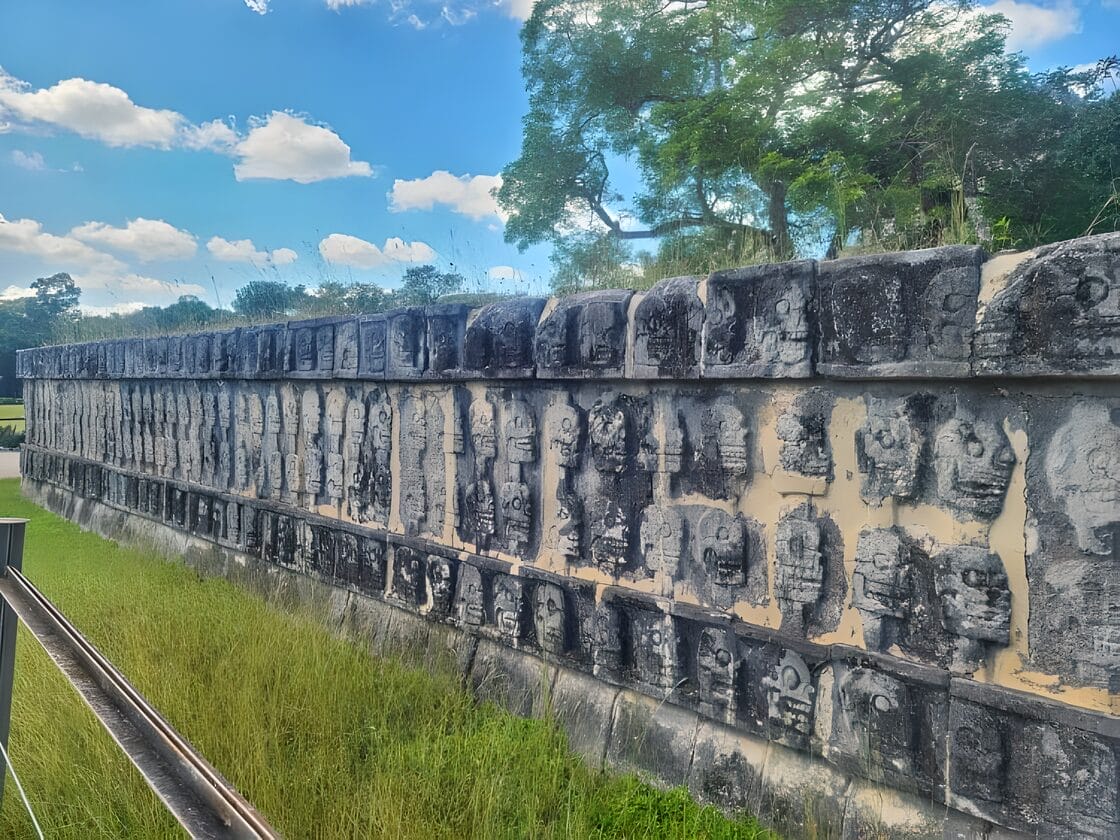 Ancient carved stone skulls on the Tzompantli (Wall of Skulls) at Chichen Itza in Yucatan, Mexico, with blue sky and greenery in the background.