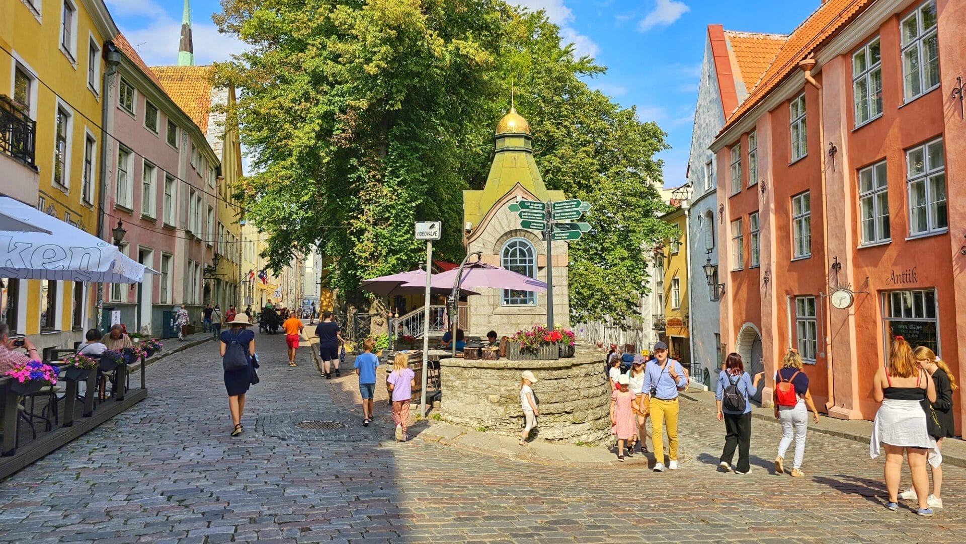 A bustling cobblestone street in Tallinn, Estonia, with pedestrians walking near colorful historic buildings, outdoor cafes, and a small chapel with a golden dome.