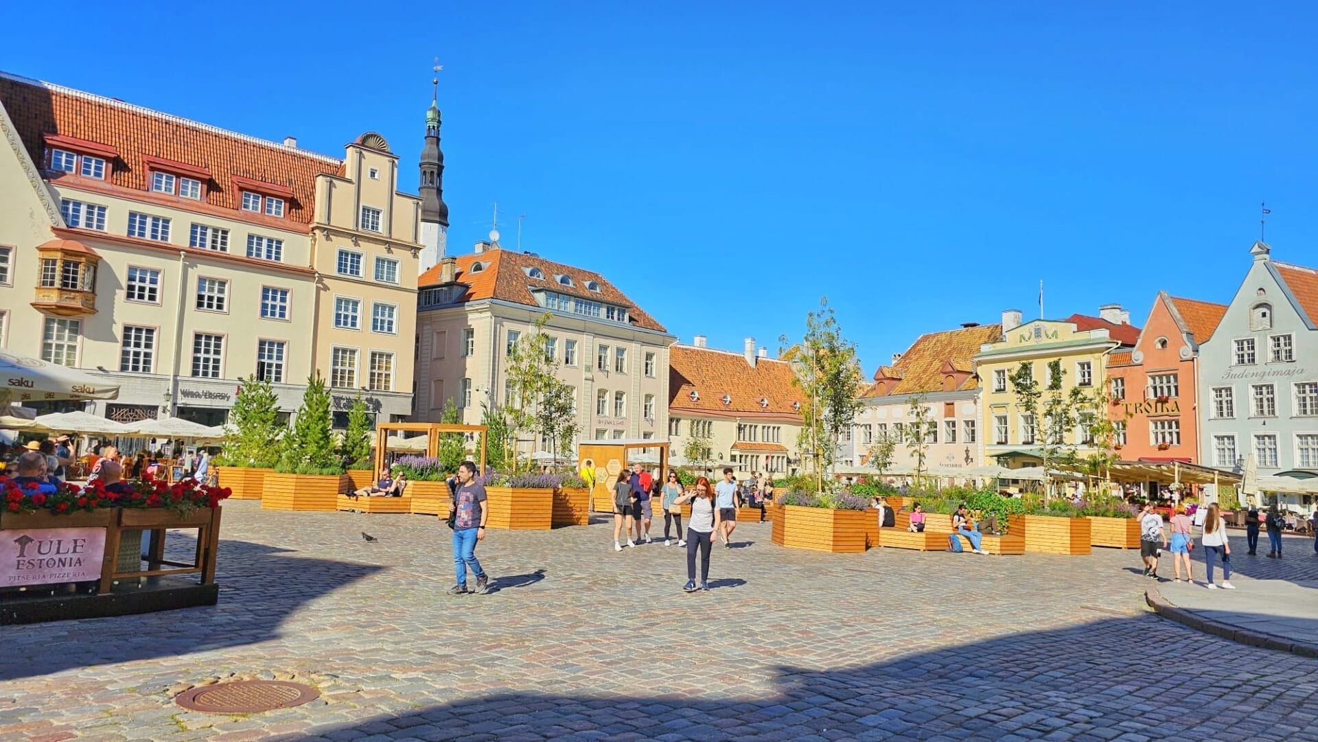 Tallinn town square with colorful historic buildings, outdoor cafes, and people walking and sitting on benches on a sunny day.