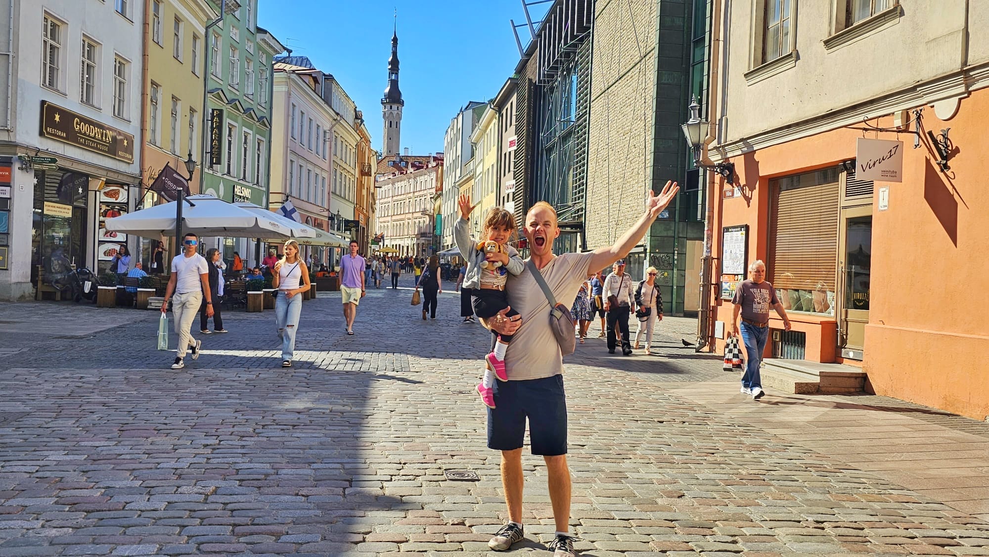 People walking on a sunny day in Old Town, Tallinn, Estonia, with colorful buildings and a man holding a child in the foreground.