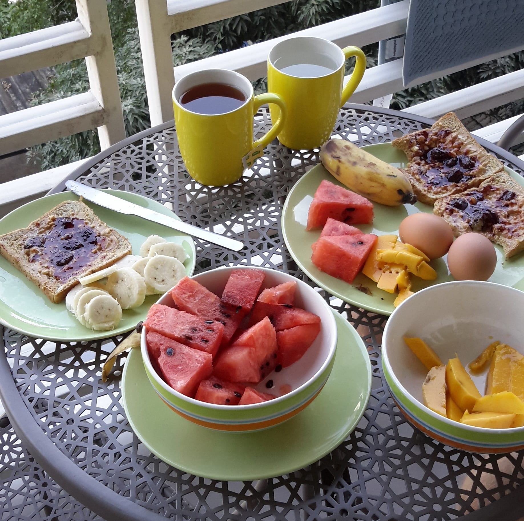 Breakfast table set with watermelon, banana, toast with jam, boiled eggs, and two cups of tea on a balcony in Chiang Mai, Thailand.