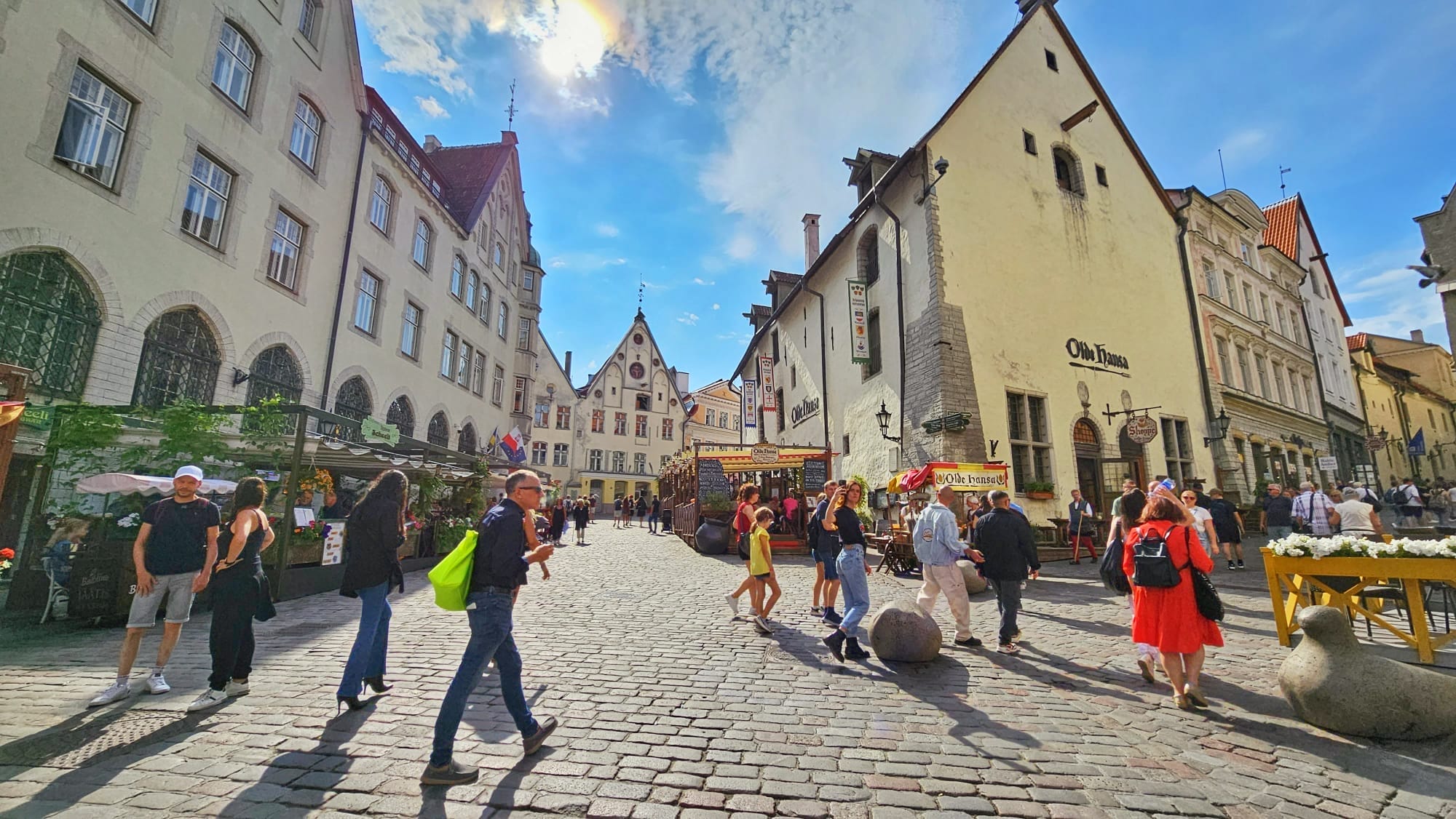 A bustling street scene in the Old Town of Tallinn, Estonia, featuring medieval buildings, people walking, and outdoor dining areas under a bright blue sky.