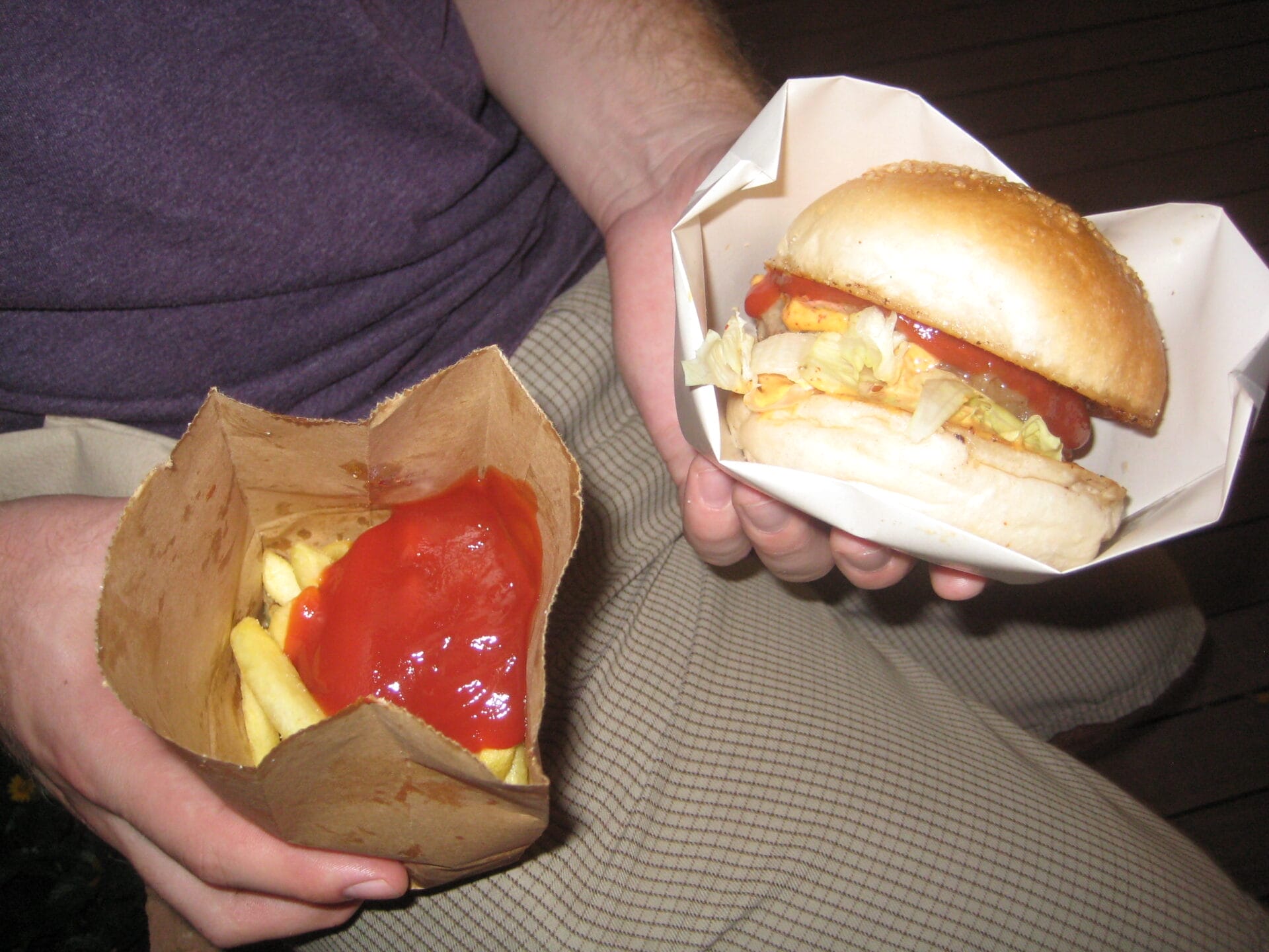 Close-up of a person holding a burger in a box with one hand and a paper bag filled with fries and ketchup in the other hand.