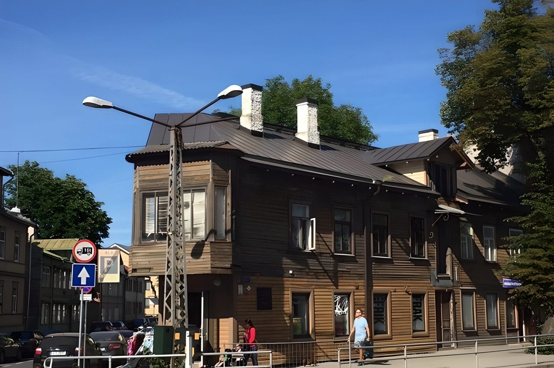 A wooden building on Kopli Street in the Kalamaja district of Tallinn, Estonia. The building features a grey metal roof with white chimneys and large windows. A few pedestrians are visible on the sidewalk, and some cars are parked nearby.