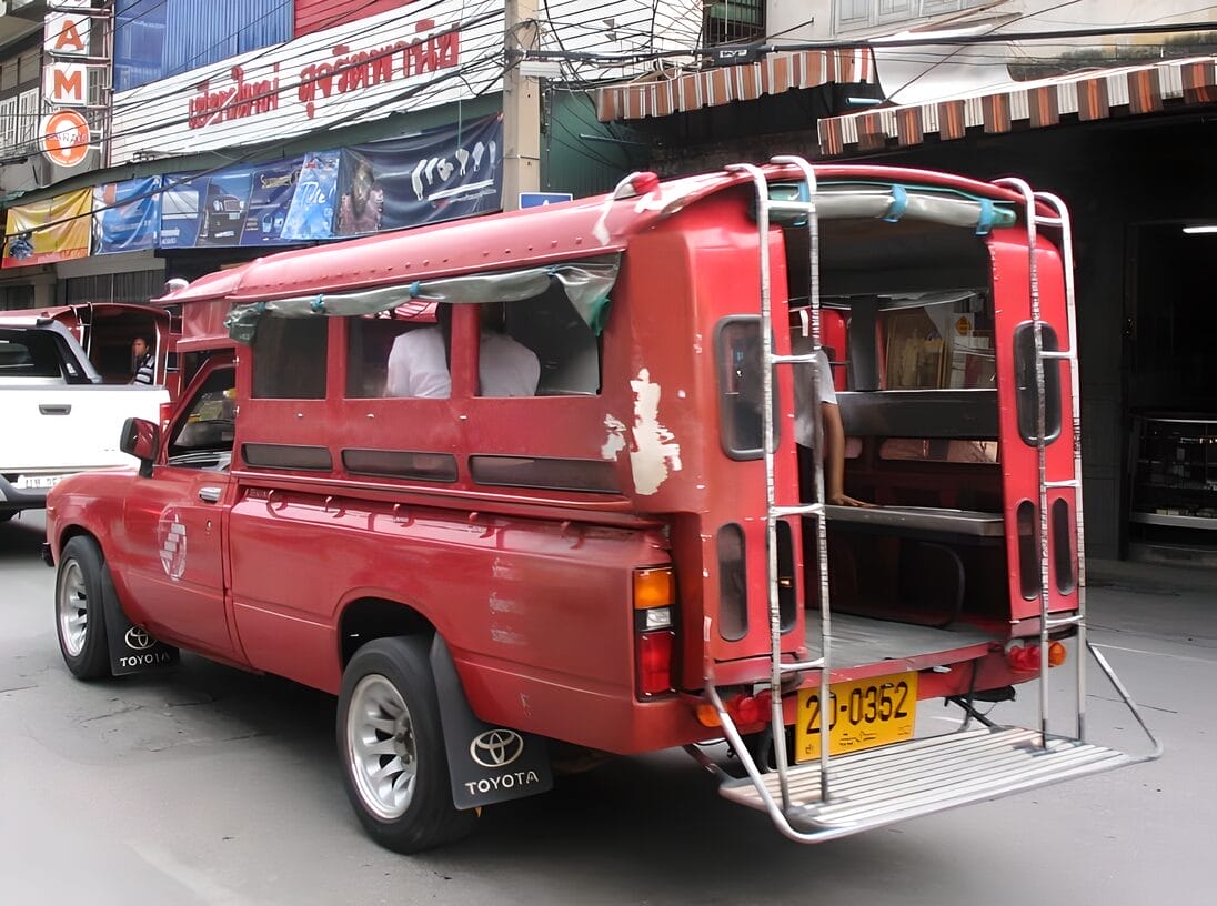 A red Songthaew bus in Chiang Mai, Thailand, parked on a city street with people seated inside.