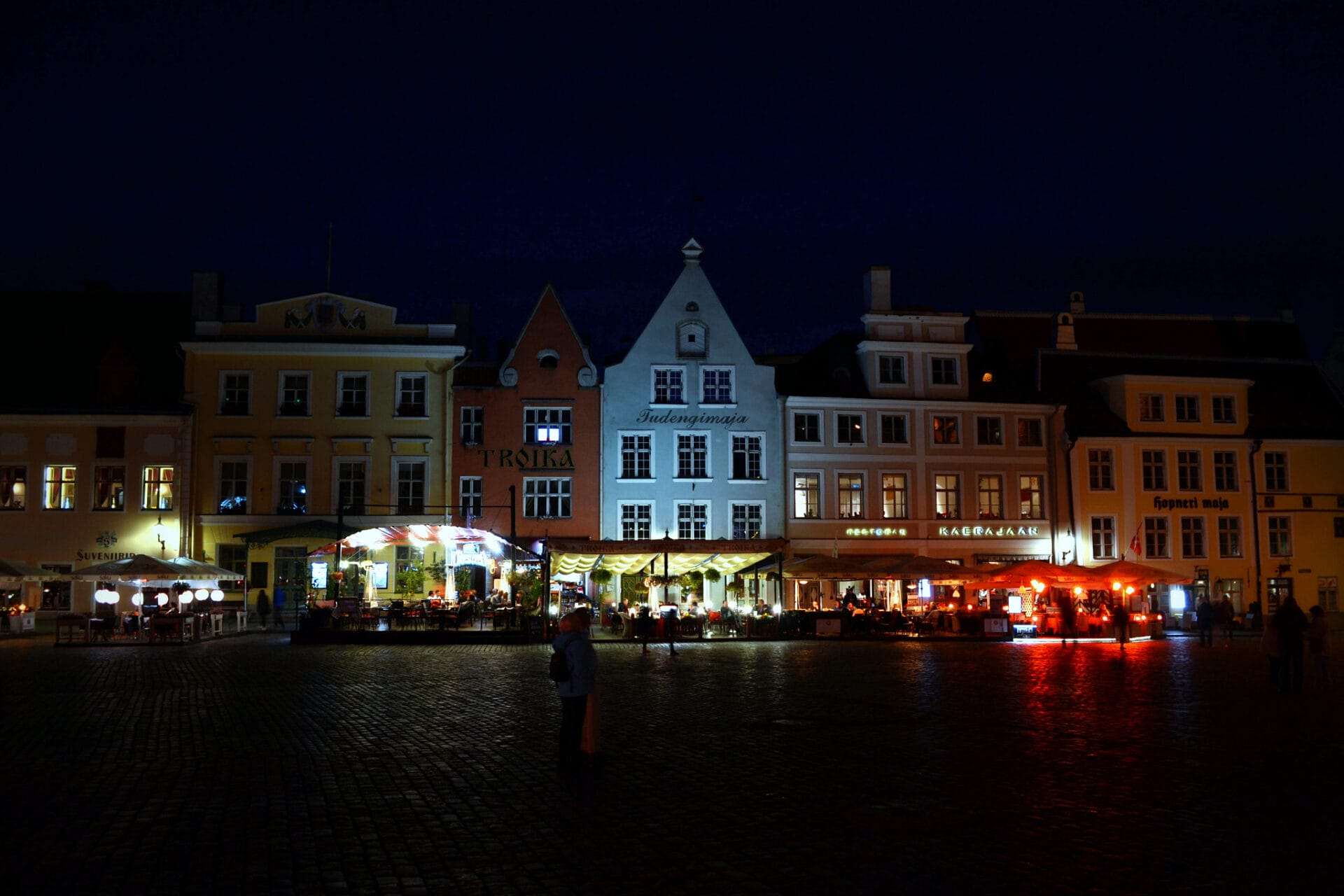 Night view of a bustling town square in Tallinn, Estonia, with illuminated buildings and outdoor cafes.
