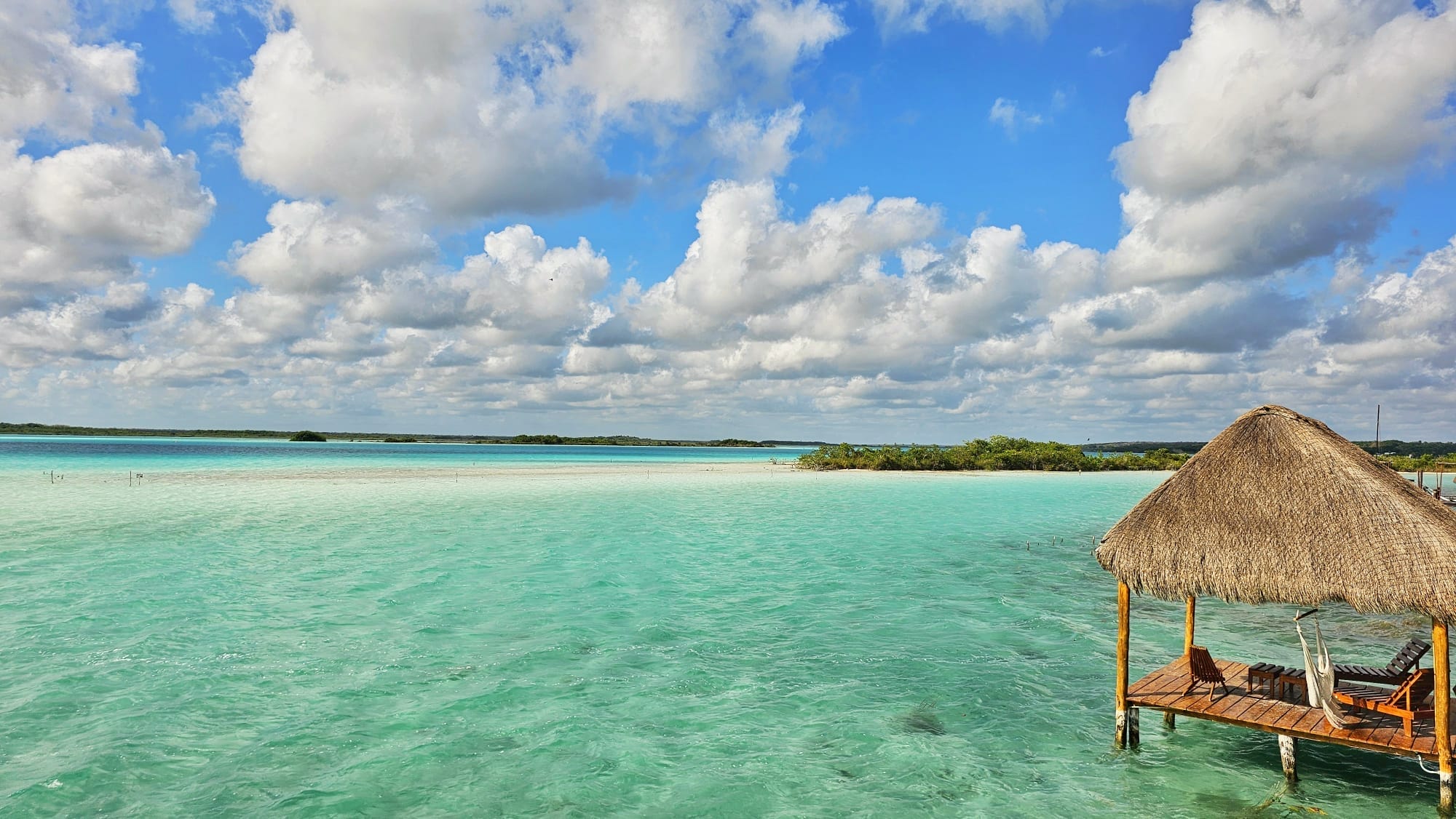 A tropical scene in Bacalar, Mexico, featuring clear turquoise waters under a partly cloudy sky. A wooden deck with a thatched roof hut extends over the water, equipped with lounge chairs and a hammock.