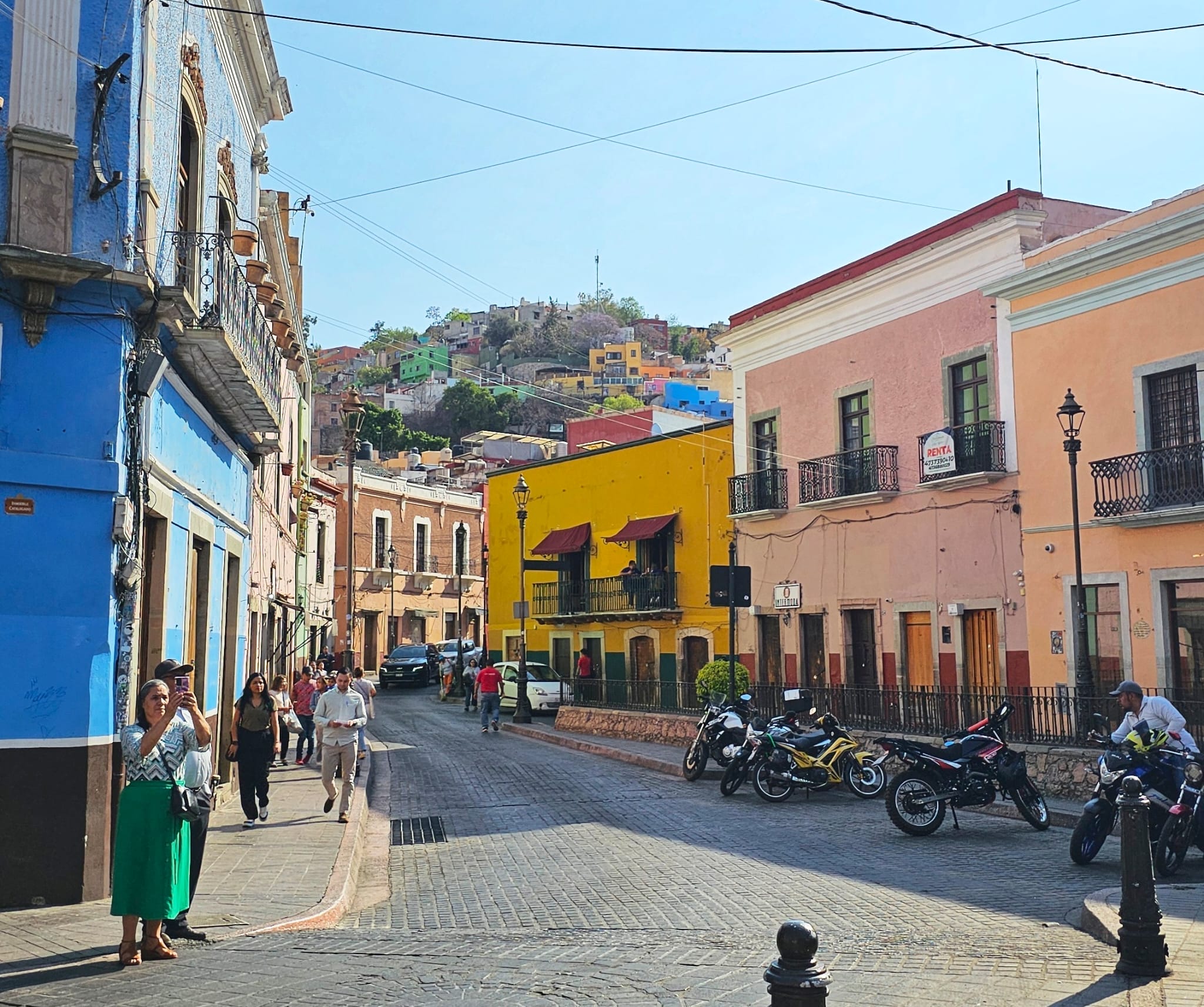 Street view in Guanajuato, Mexico, showcasing colorful colonial buildings and people walking, with a hill and houses in the background.