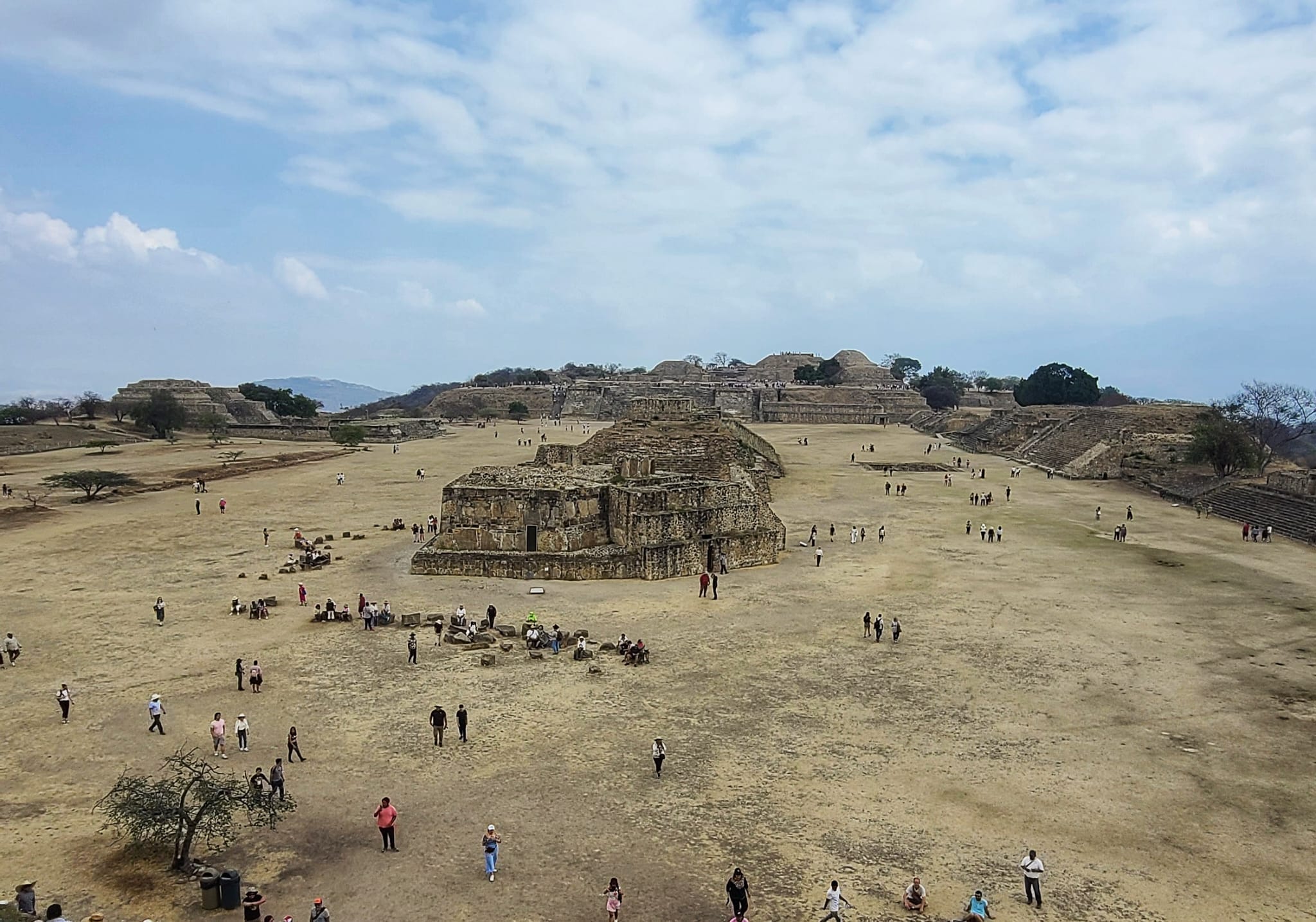 Aerial view of the ancient ruins at Monte Alban in Oaxaca, Mexico, with numerous visitors exploring the historical site under a partly cloudy sky.