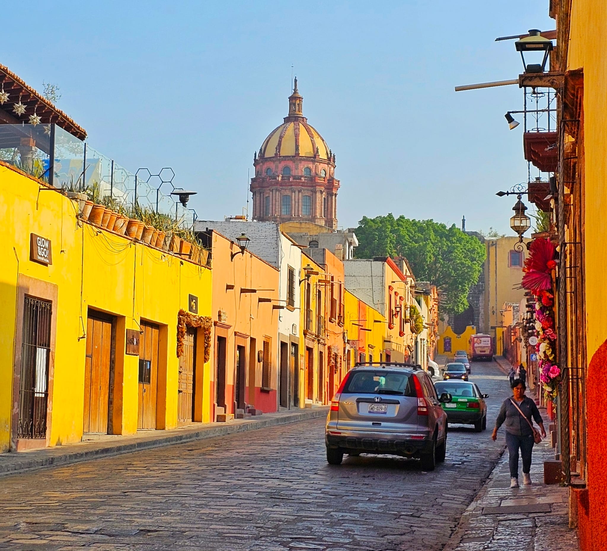 A cobblestone street in San Miguel de Allende, Mexico, lined with colorful buildings. In the background, the dome of a historic church rises above the rooftops. A few cars are parked along the street, and a person is walking on the sidewalk.