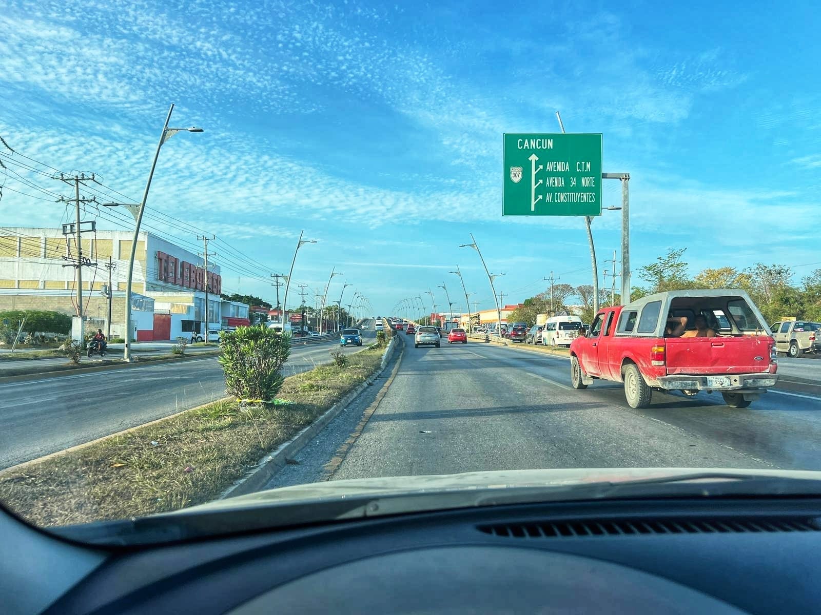 A busy highway in Quintana Roo, Mexico with a sign indicating directions to Cancun. Various vehicles, including a red pickup truck, are traveling in both directions. 