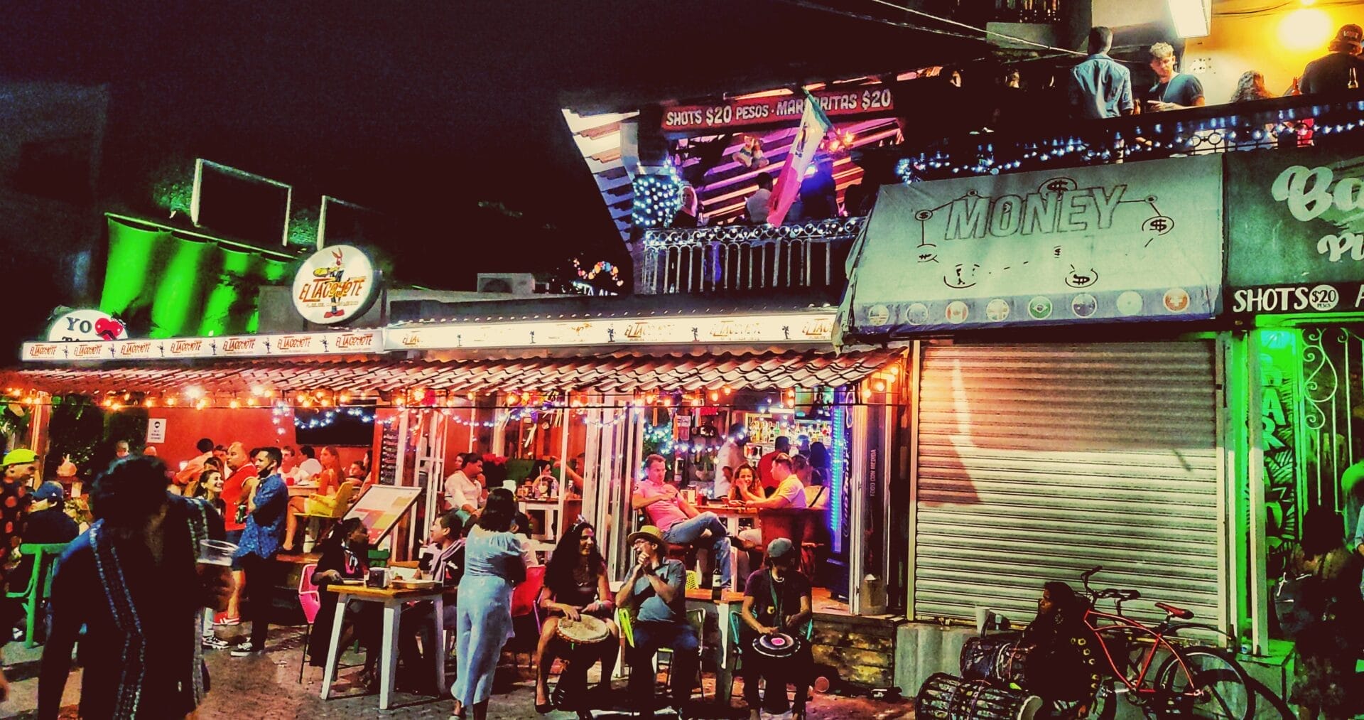 Bustling nightlife scene with people socializing at a colorful outdoor bar in Playa del Carmen, Riviera Maya, with visible signage offering drink specials.